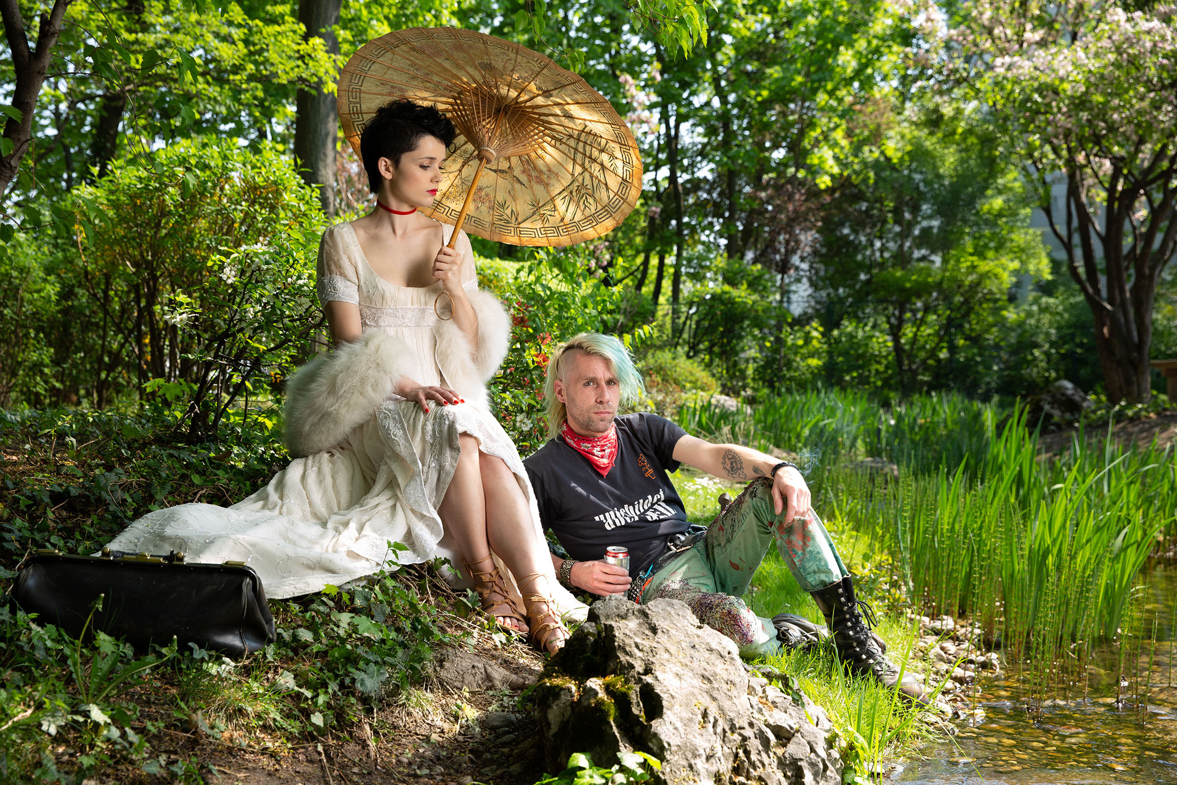 1800s fashion inspired advertising photoshoot in setagaya park by fashion historian and stylist katalina sasdy. a lady in a white empire dress with a white fur stole and a red velvet collar, wearing greek shoes, with an umbrella in her hands, is sitting with a punk by a stream. she is wearing the so called titus hairstyle, which means that the hair is shaved at the back and short curls are placed at the front. the punk is wearing a funny t shirt, a red small scarf, ripped jeans and biker boots. his green hair is shaved on the side and sticks up in the middle.