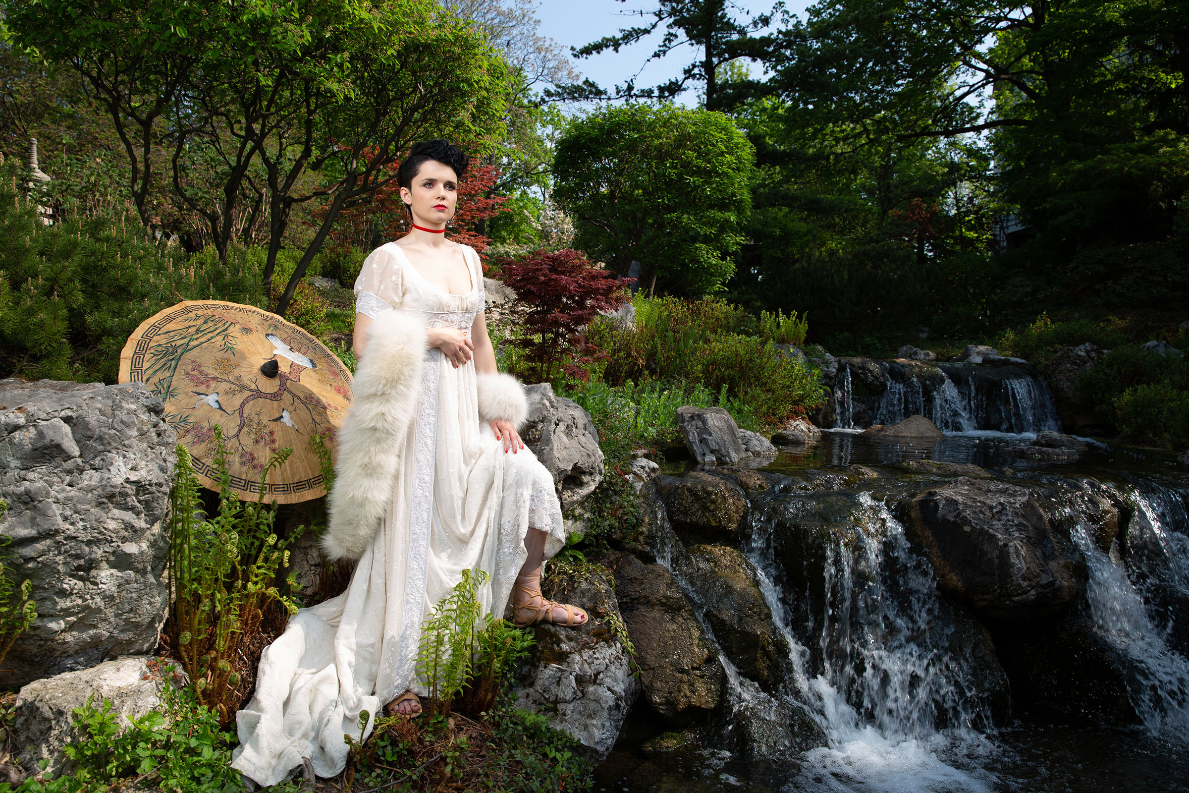 fashion of the directoire inspired advertising photo shoot in setagaya park by fashion stylist katalina sasdy. a lady in a white empire dress and white fur stole with a red velvet collar is standing next to a small waterfall. behind her a japanese umbrella is located. she places her bare feet on a rock, revealing her beige greek sandals. she is wearing the so called titus hairstyle, which means that the hair is shaved at the back and short curls are placed at the front.