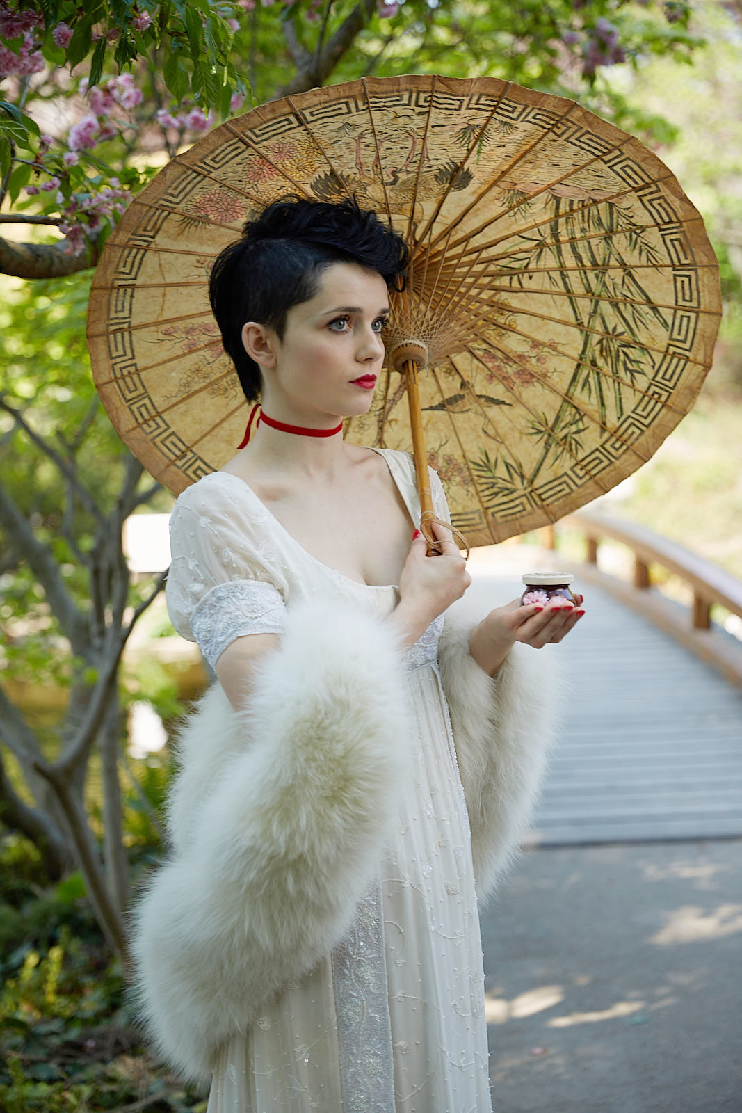 age of napoleon inspired darbo advertisng photo shoot in setagaya park by fashion stylist katalina sasdy. a lady in a white empire dress with a white fur stole and a red velvet collar, wearing greek shoes, with an umbrella in her hands, is sitting on a stone, in the garden. she is wearing the so called titus hairstyle, which means that the hair is shaved at the back and short curls are placed at the front. she is holding d arbo fruchtikus, decorated with white flowers in her hand. behind her are some green trees and bushes.