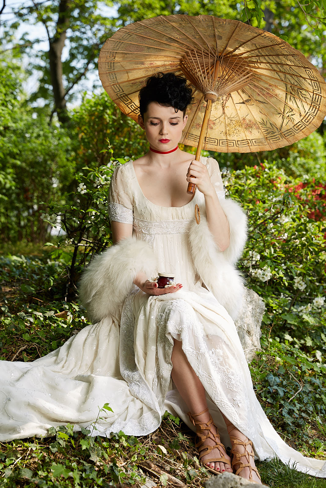 bridgerton by netflix inspired advertising photoshoot in setagaya park by art director and fashion stylist katalina sasdy. a lady in a white empire dress with a white fur stole and a red velvet collar, wearing greek shoes, with an umbrella in her hands, is sitting with a punk by a stream. she is wearing the so called titus hairstyle, which means that the hair is shaved at the back and short curls are placed at the front. the punk is wearing a a funny t shirt, a red small scarf, ripped jeans and biker boots. his green hair is shaved on the side and sticks up in the middle. he is looking at her lovingly.