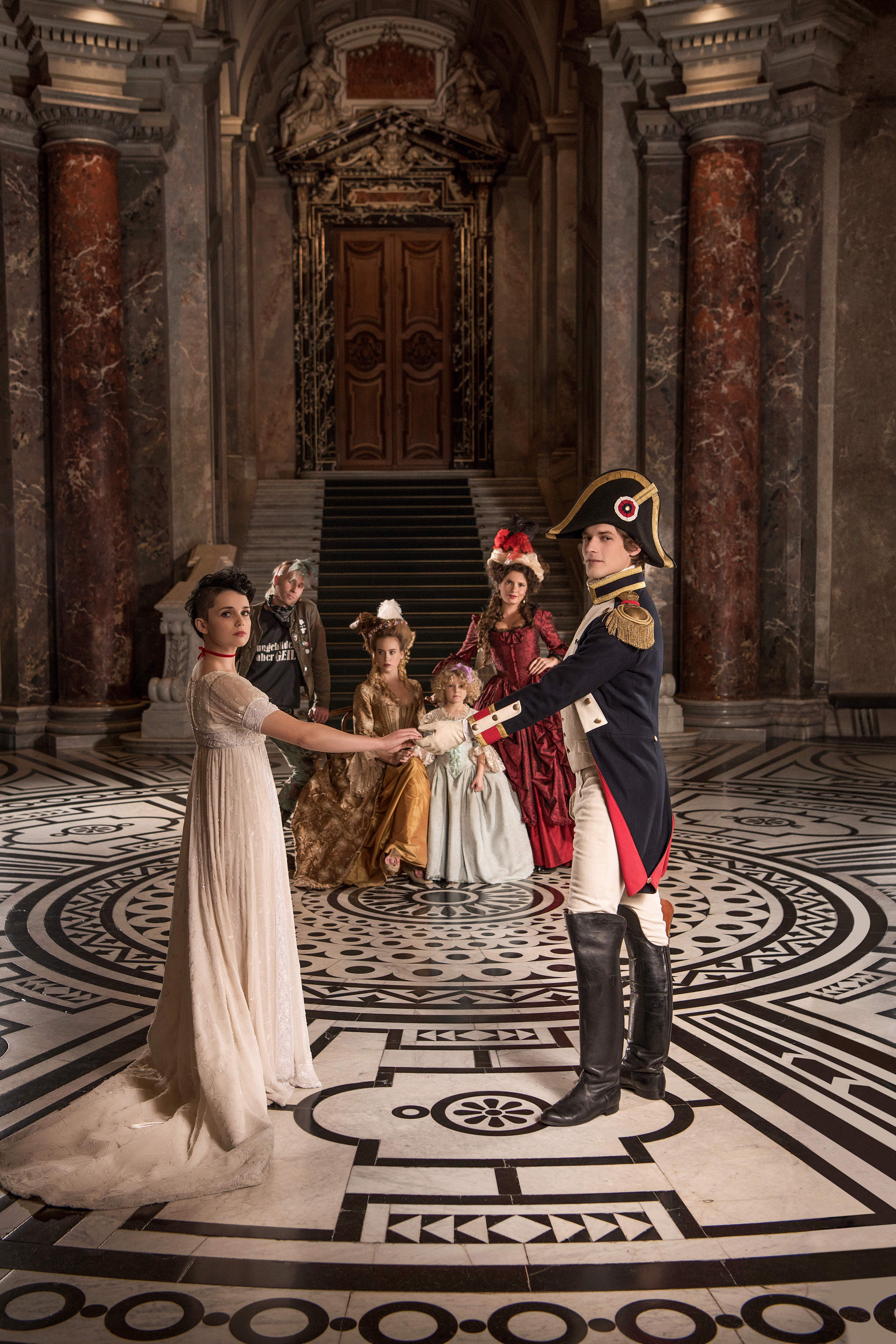 rokoko and regency fashion inspired advertising photo shoot in khm vienna by fashion stylist katalina sasdy. there are four people in the background, two ladies, each in yellow and red rococo dresses, a little girl in a light blue rococo dress and a punk in a provocative outfit with raised green hair. a soldier in the uniform of the napoleonic army is standing in front of the group and holding the hand of a lady in a white empire dress and black shaved hair.