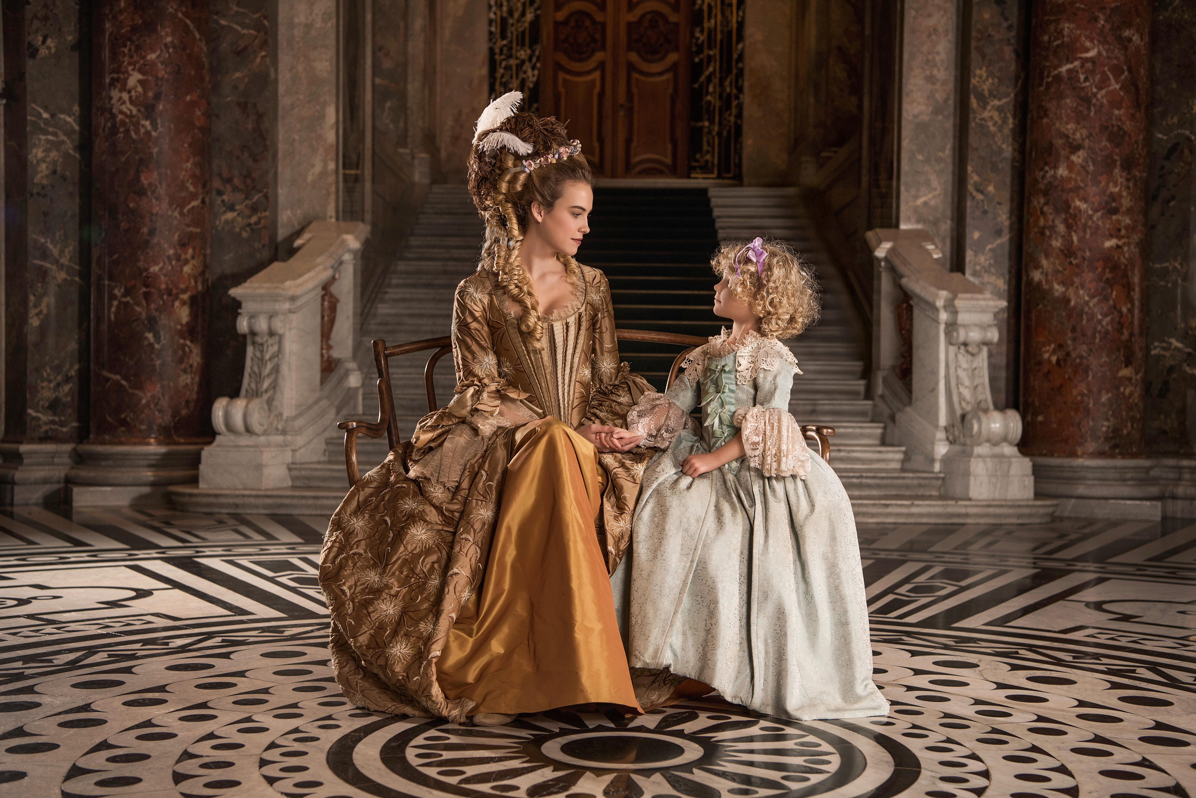 marie antoinette inspired advertising photo shoot in khm vienna by fashion stylist katalina sasdy. marie antoinette is sitting on a sofa in a yellow silk dress with her little daughter marie therese. mother and daughter are looking at each other in love and holding hands.