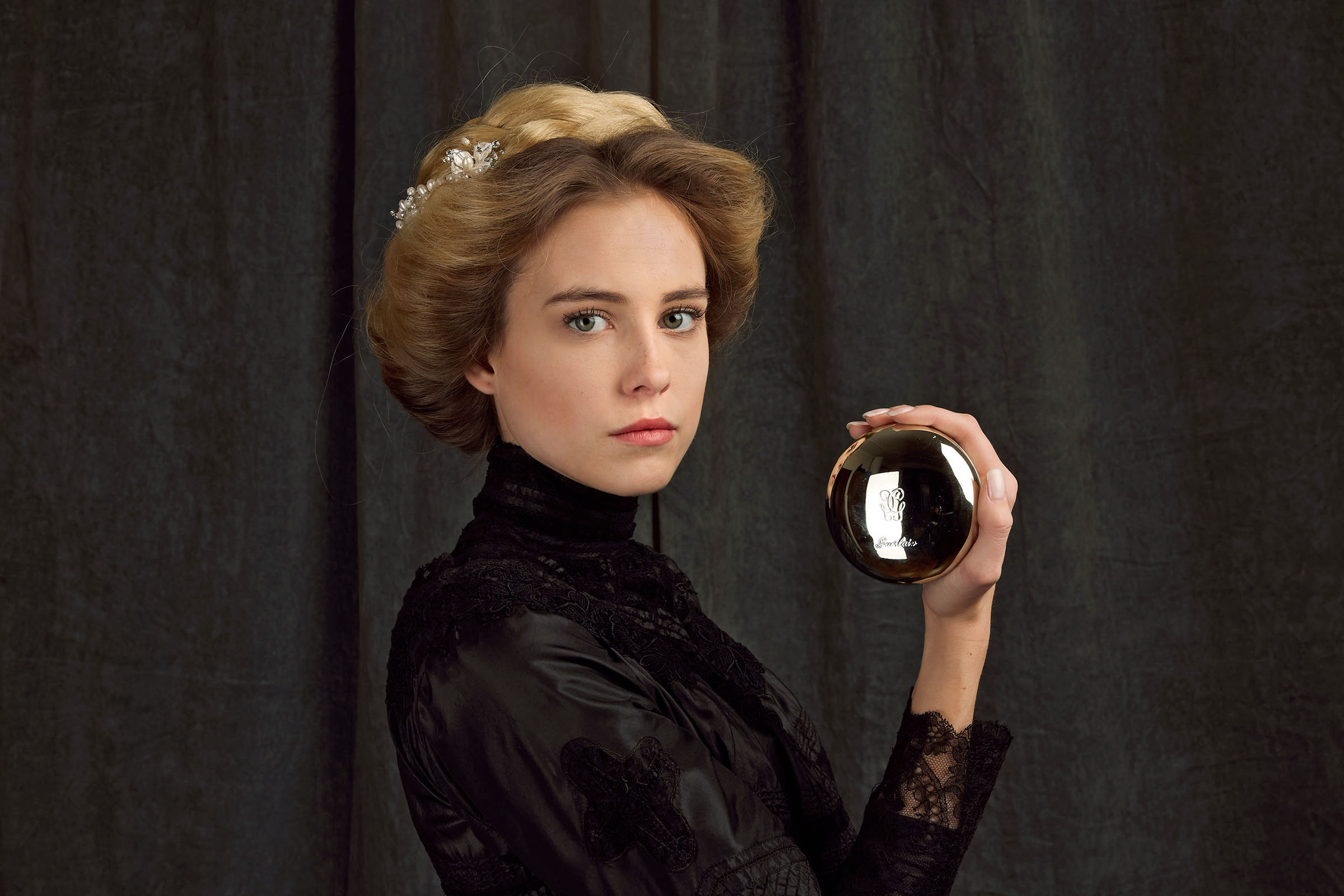 19th century fashion inspired guerlain advertising photo shoot by fashion historian and stylist katalina sasdy. a blonde lady in a black, 1900s style silk and lace blouse, is holding a silver powder compact by guerlain in her hand and is looking into the camera. her hairstyle is pinned up in the style of the belle epoque and held together with a silver hair clip with pearls.