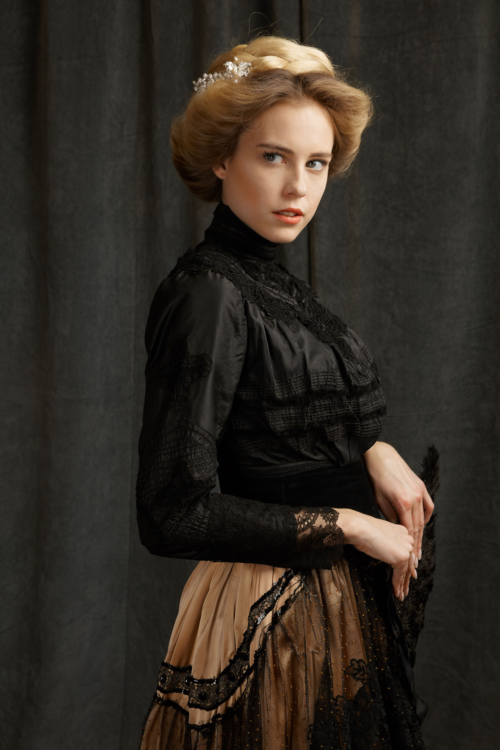 19th century fashion inspired advertising photo shoot by fashion stylist katalina sasdy. a blonde lady in a black, 1900s style silk and lace blouse and a pink silk skirt with black lace, including a black belt, is holding a black strauss feather fan in her hands and is looking flirty into the camera. her hairstyle is pinned up in the style of the belle epoque and held together with a silver hair clip with pearls.
