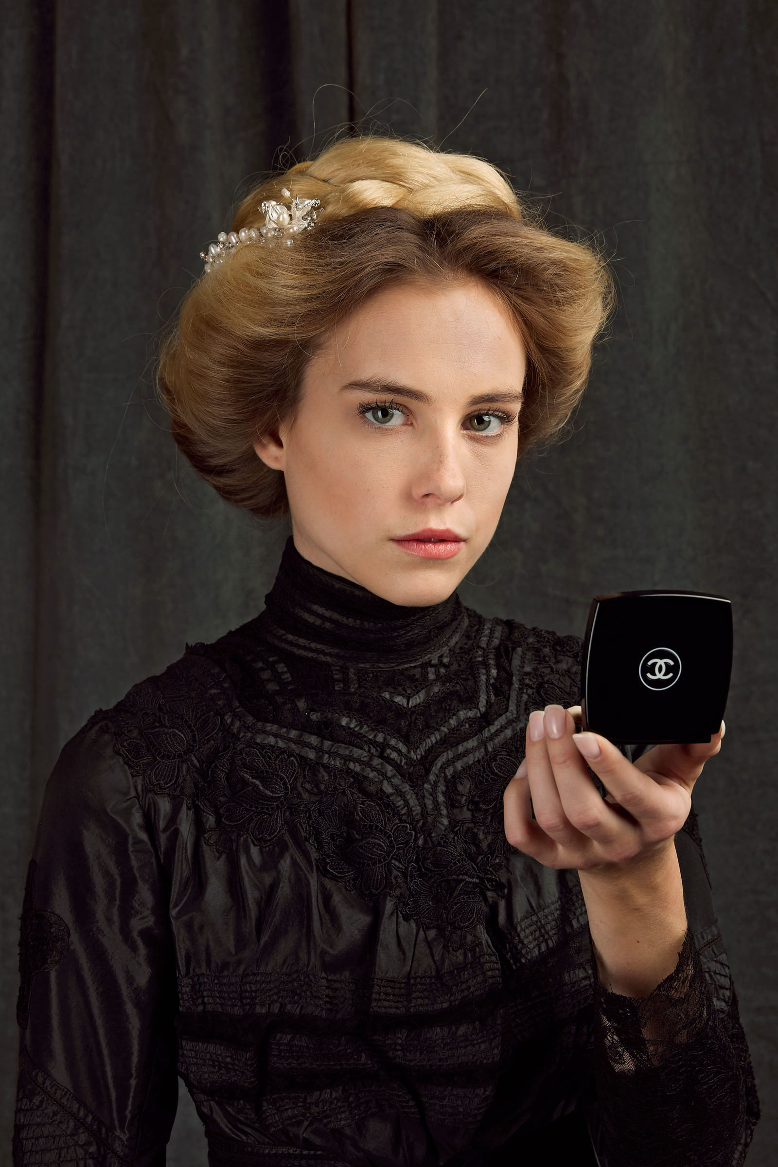 19th century fashion inspired chanel advertising photo shoot by art director and fashion stylist katalina sasdy. a blonde lady in a black, 1900s style silk and lace blouse, is holding a black powder compact from chanel in her hand and is looking into the camera. her hairstyle is pinned up in the style of the belle epoque and held together with a silver hair clip with pearls.
