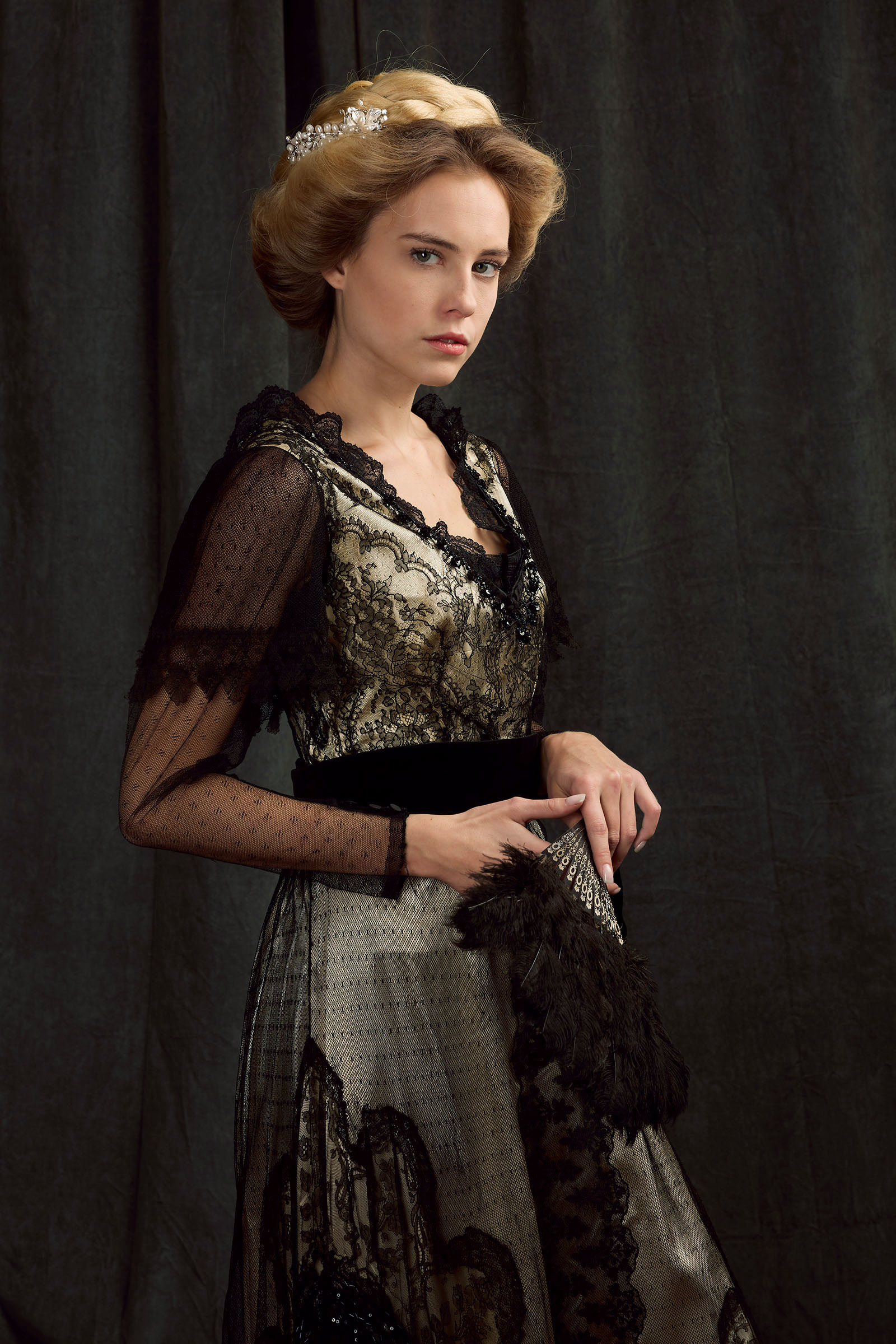 belle epoque fashion inspired advertising photoshoot by fashion stylist katalina sasdy. a blonde lady in a creme 1900s style silk dress with black lace is holding a black strauss feather fan in her hand and is looking flirty into the camera. her hairstyle is pinned up in the style of the belle epoque and held together with a silver hair clip with pearls.