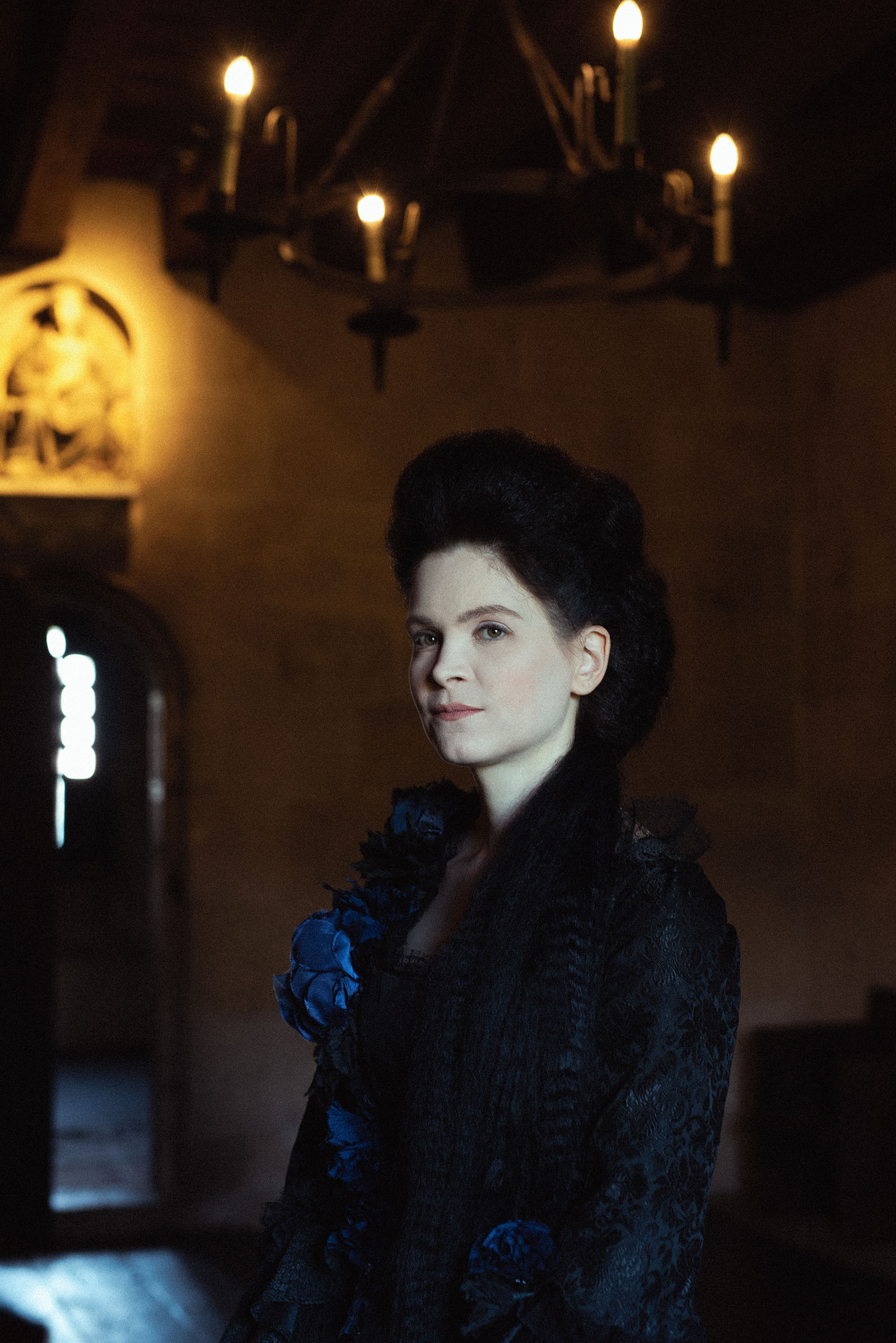 vampire inspired advertising photo shoot at the castle of liechtenstein by art director and fashion stylist katalina sasdy. a lady with pinned black hair and a black rococo dress is standing in an illuminated room. the door is half open. a medieval chandelier is hanging above her.