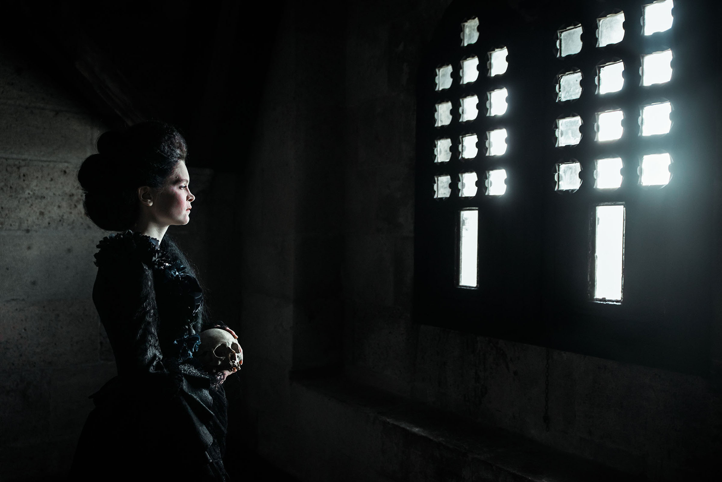 18th century inspired halloween advertising photo shoot at the castle of liechtenstein by fashion historian and stylist katalina sasdy. a lady with pinned black hair and a black rococo dress is standing in a dark room that only gets light from the window grilles. in her hands she is holding a skull.