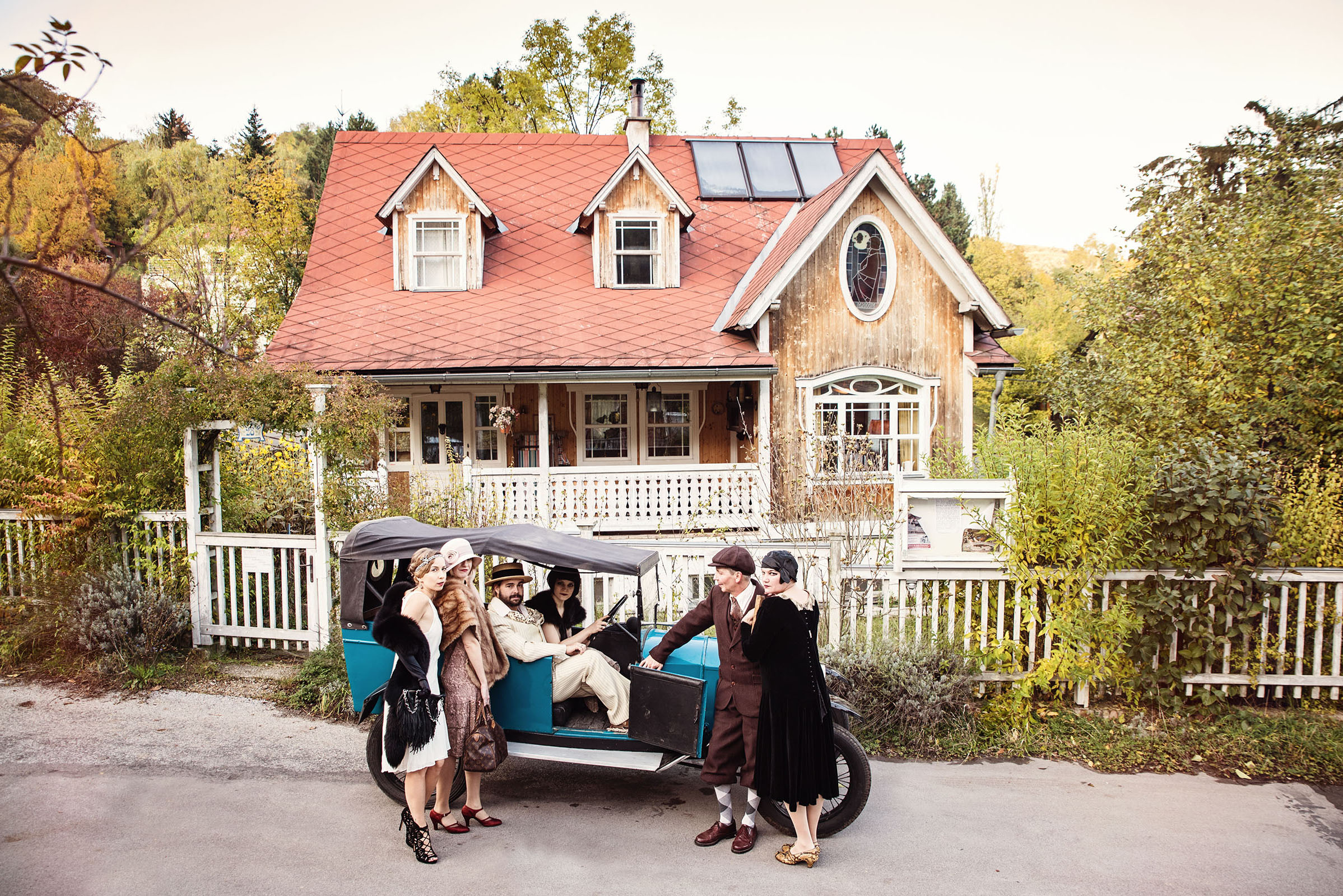 1920s fashion inspired advertising photography with oldtimer by fashion historian and vintage stylist katalina sasdy. a group of people, dressed in 1920s fashion by lambert hofer standing in front of a house from the 1920s. on the road is an oldtimer as well, which is sorrounded by the people. one of the ladies is driving the car.