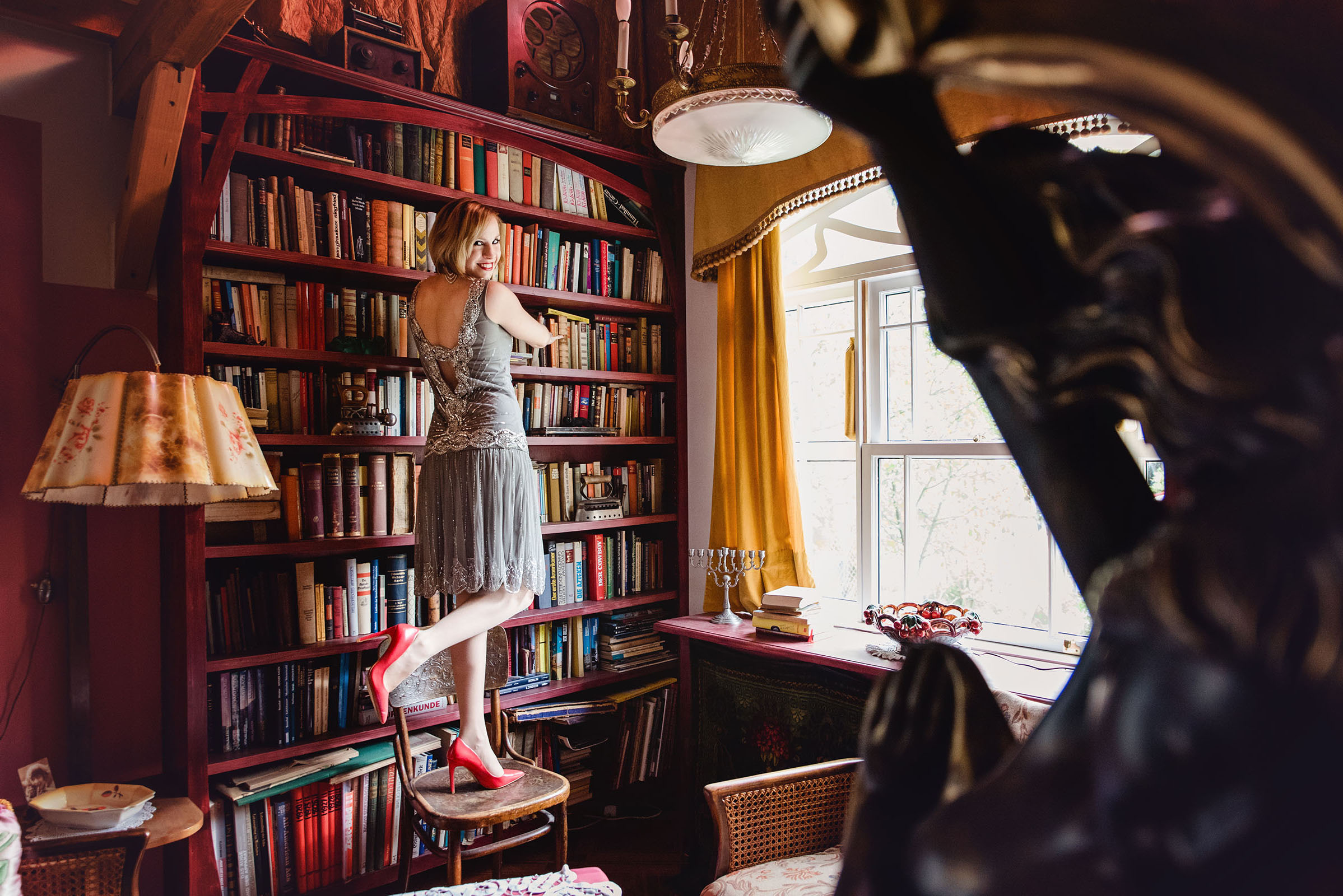 the great gatsby themed frock and frill advertising photo shoot by art director and vintage stylist katalina sasdy. a lady is standing with her leg raised on an armchair in front of a huge, colorful bookshelf. she is wearing a grey frock and frill chiffon dress and red leather shoes by zara.