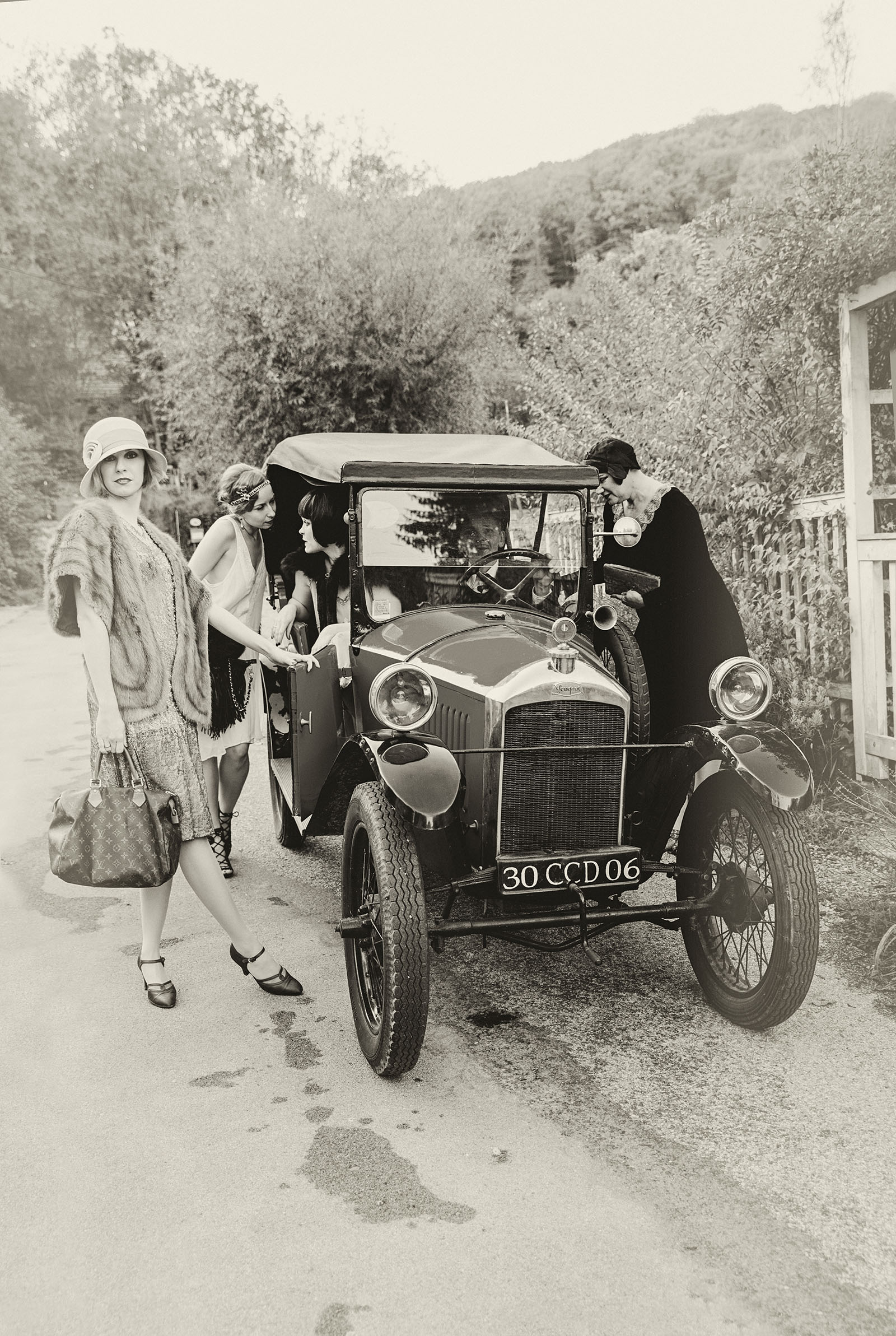 the great gatsby inspired advertising photo shoot with oldtimer by art director and vintage stylist katalina sasdy. three ladies are standing around a vintage car. the fourth of the women is driving the car. everyone is dressed in the style of the 1920s. one of the ladies is holding in her hand a louis vuitton bag. the other two ladies are leaning towards the car and talking to the driver.
