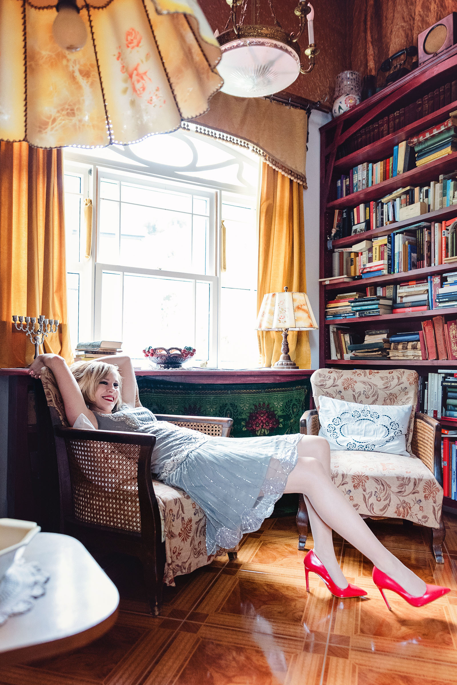 the great gatsby themed frock and frill advertising photoshoot by fashion historian and vintage stylist katalina sasdy. a lady is stretching out comfortably on an armchair. she is wearing a grey chiffon frock and frill dress and red shoes by zara.