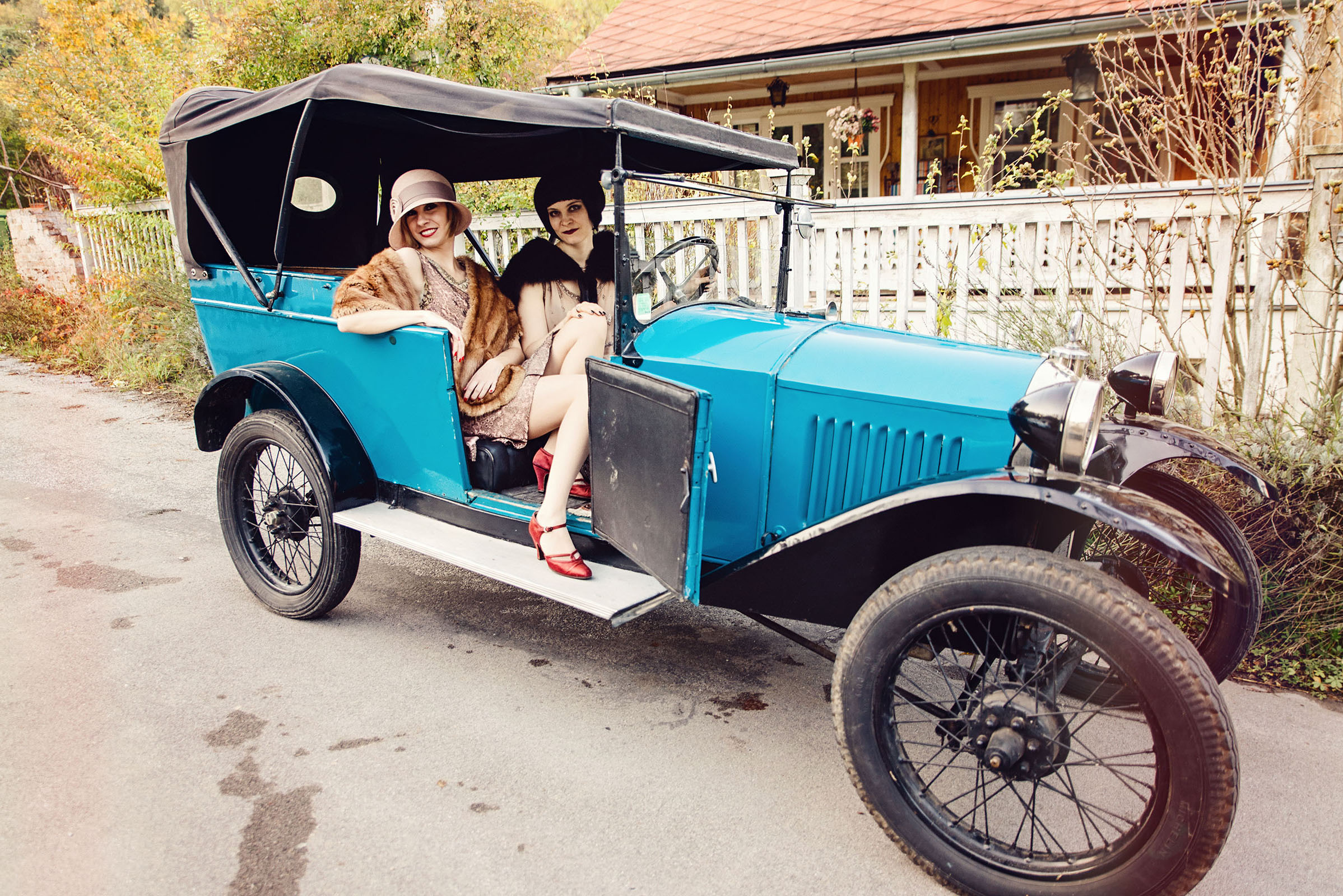 1920s fashion inspired advertising photography with oldtimer by vintage fashion stylist katalina sasdy. two ladies are sitting in a blue vintage car. both women are wearing 1920s fashion. one with black hair is wearing a light pink dress with a black fur stole. the other lady, with blonde hair, is also wearing a light pink dress with a beige fur stole and a pink straw hat including red leather shoes.