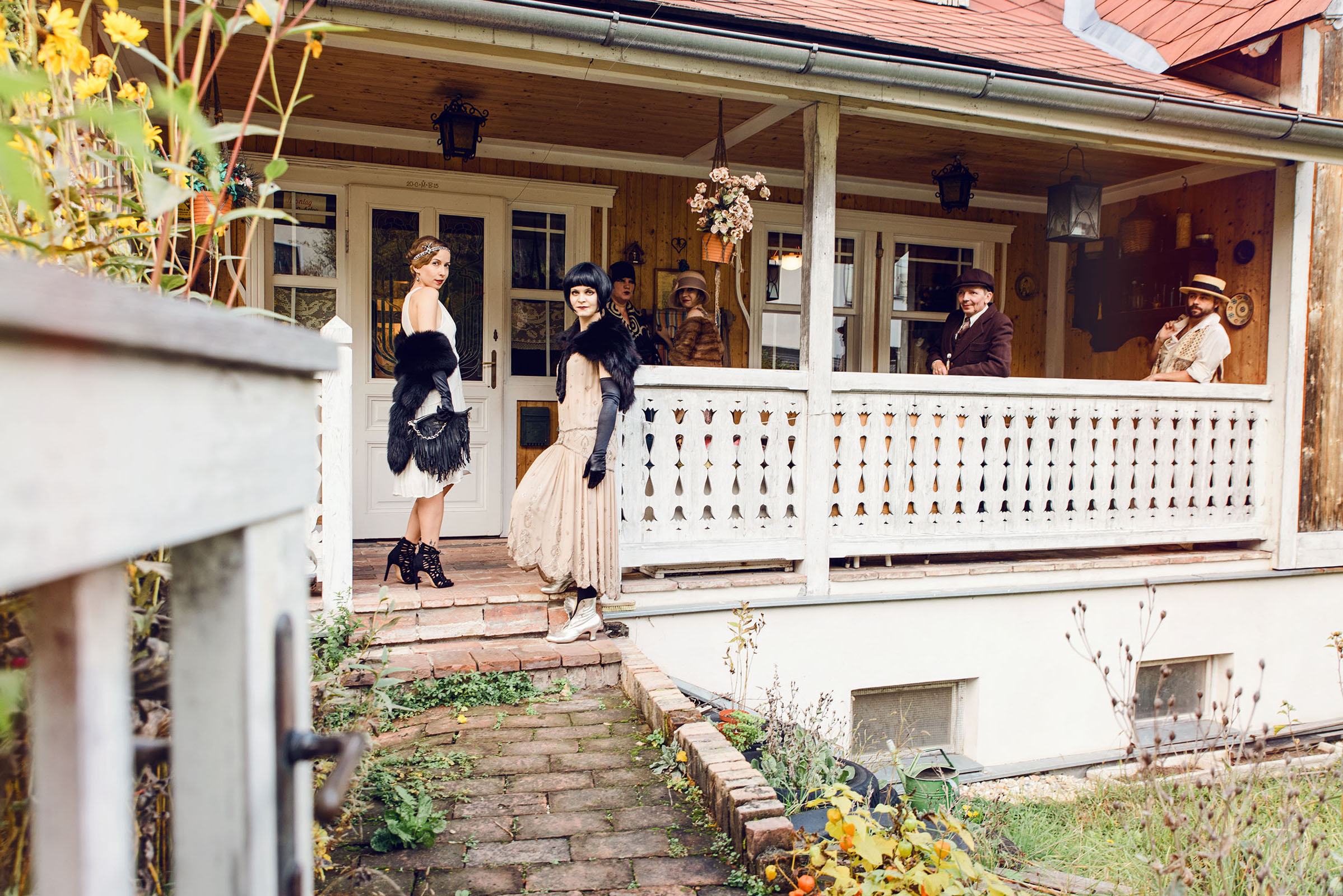 1920s womens fashion inspired advertising photo shoot by art director and vintage stylist katalina sasdy. the white garden gate is open and it is possible to see the whole group on the terrace. two ladies are standing on the stairs in white and light pink dresses with black fur stoles and black gloves. they are wearing black high heels by zara and original 1920s silver boots.