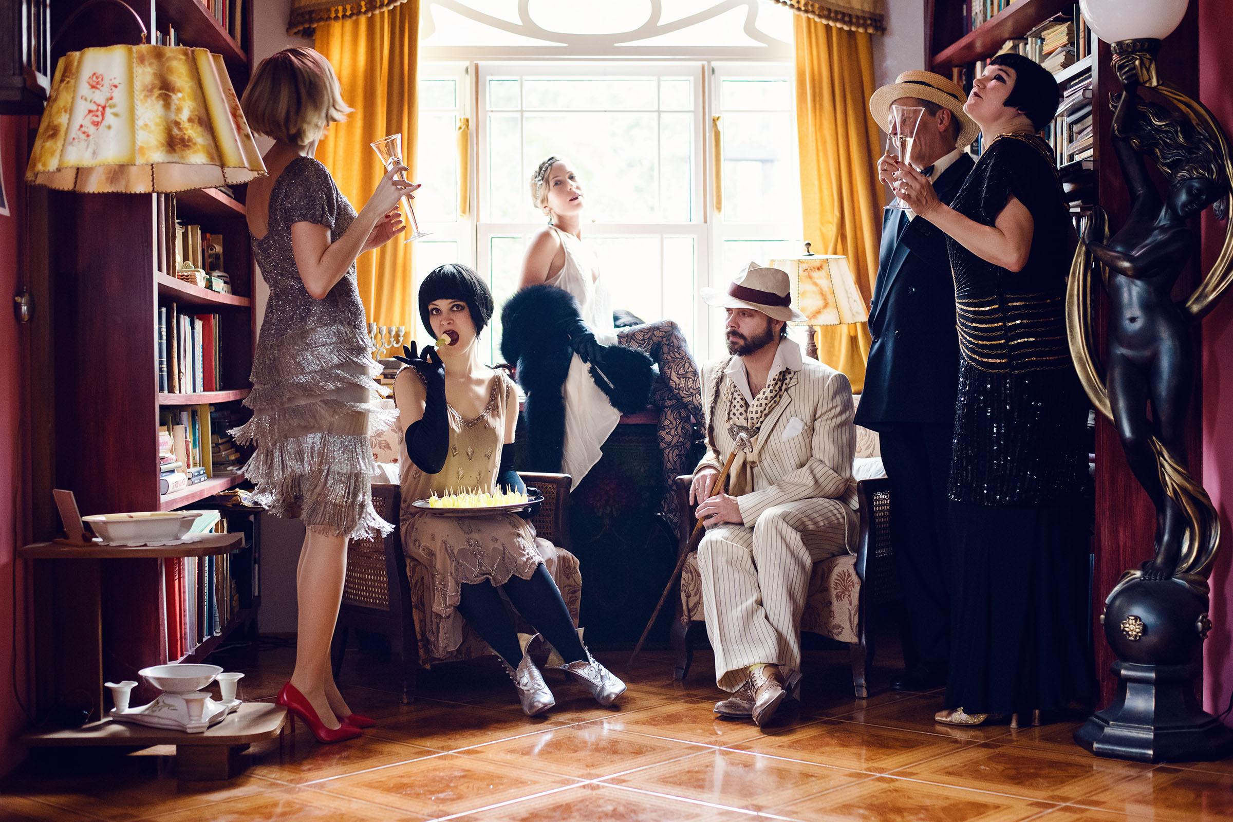 the great gatsby inspired wedding party photo shoot by fashion historian and vintage stylist katalina sasdy. a group of six people toasting a party with champagne glasses. everyone is dressed in the style of the 1920s. the ladies are wearing white, light pink, silver and black evening dresses with gloves and fur stoles. the gentlemen are wearing creme and black suits with beige hat and black walking stick.