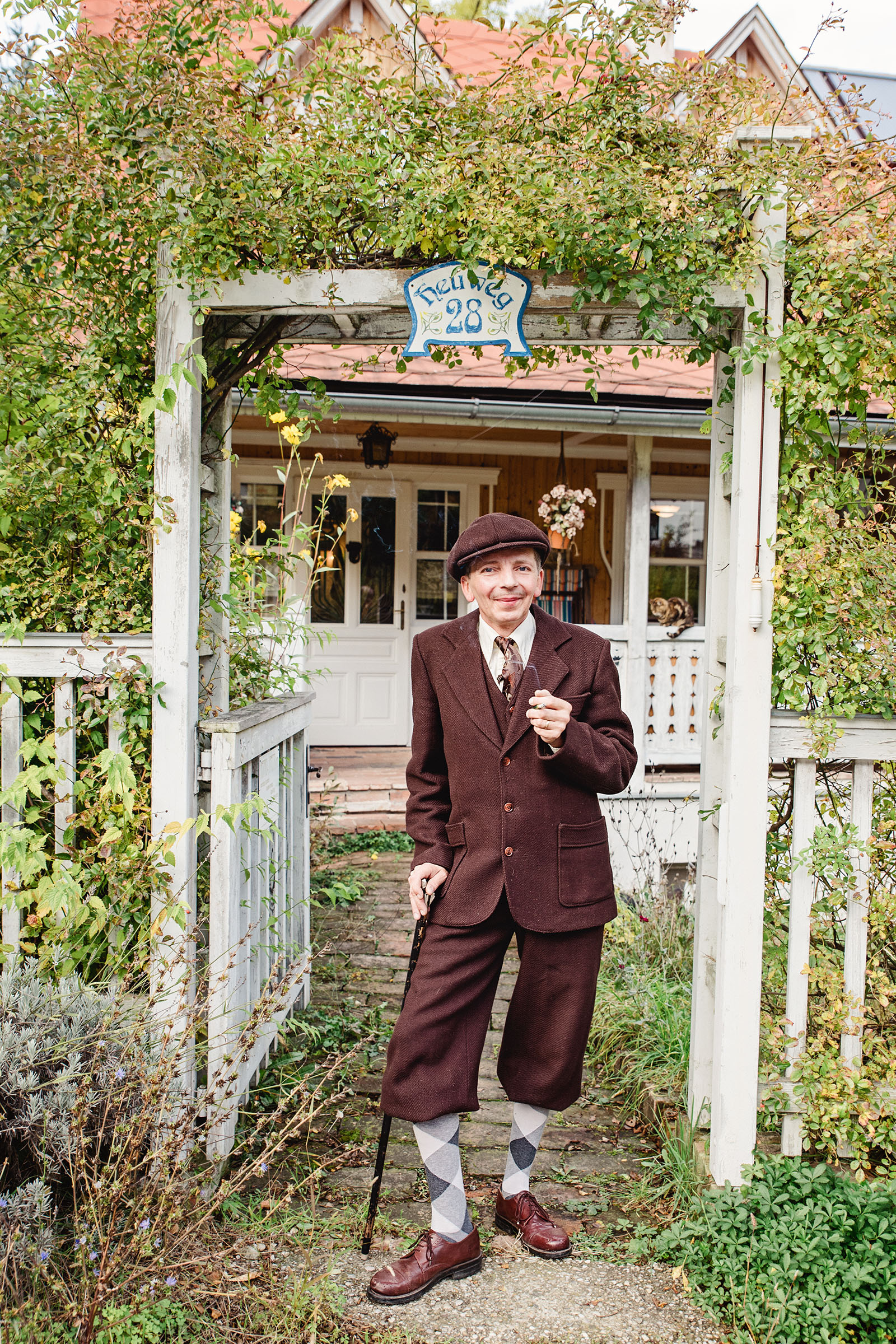 1920s mens fashion inspired advertising photo shoot by vintage fashion stylist katalina sasdy. a gentleman in a brown 1920s look is standing in front of the white garden gate. he is wearing checkered long socks, a brown cap and is leaning on a walking stick.