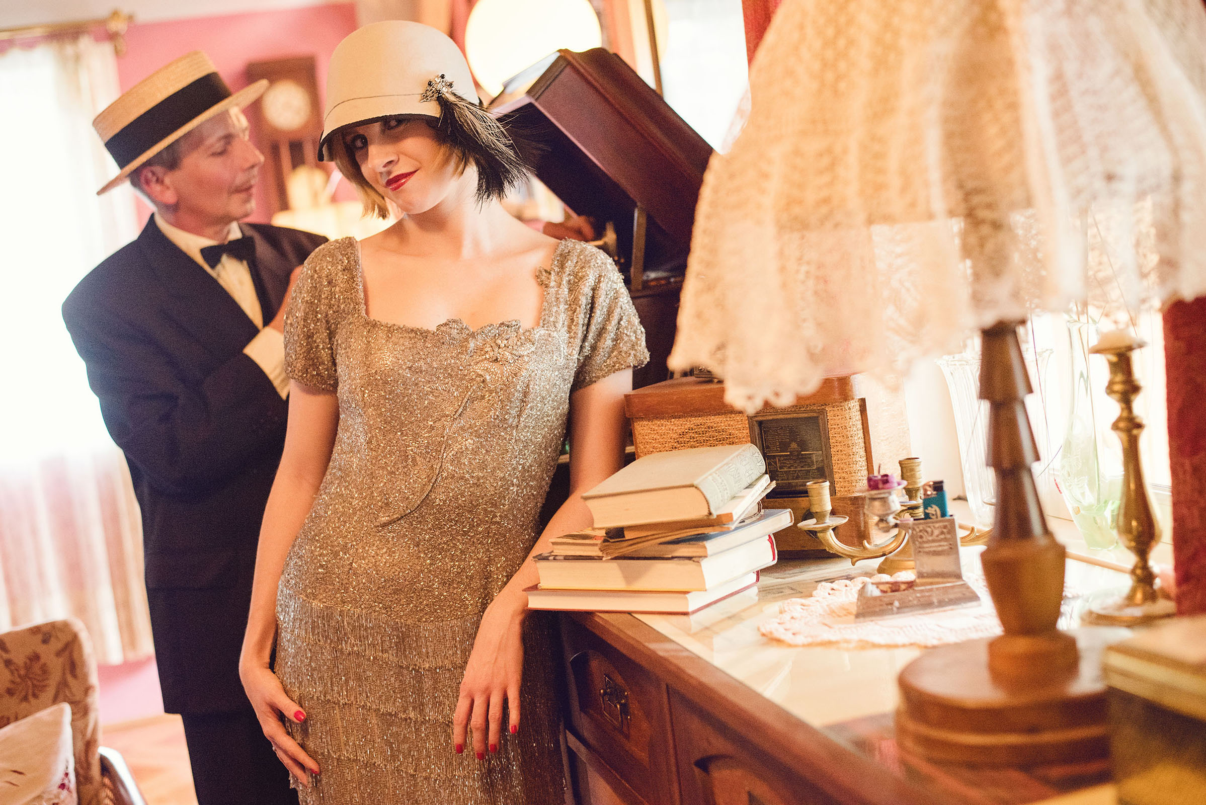 1920s inspired wedding photo shoot by art director and vintage stylist katalina sasdy. a lady is leaning against a dresser in a silver beaded dress, wearing a light grey hat with black feathers. behind her, a gentleman, wearing a black suit and a straw hat, is playing a record on a gramophone. on the dresser is a lamp, books, an old radio and a candlestick.