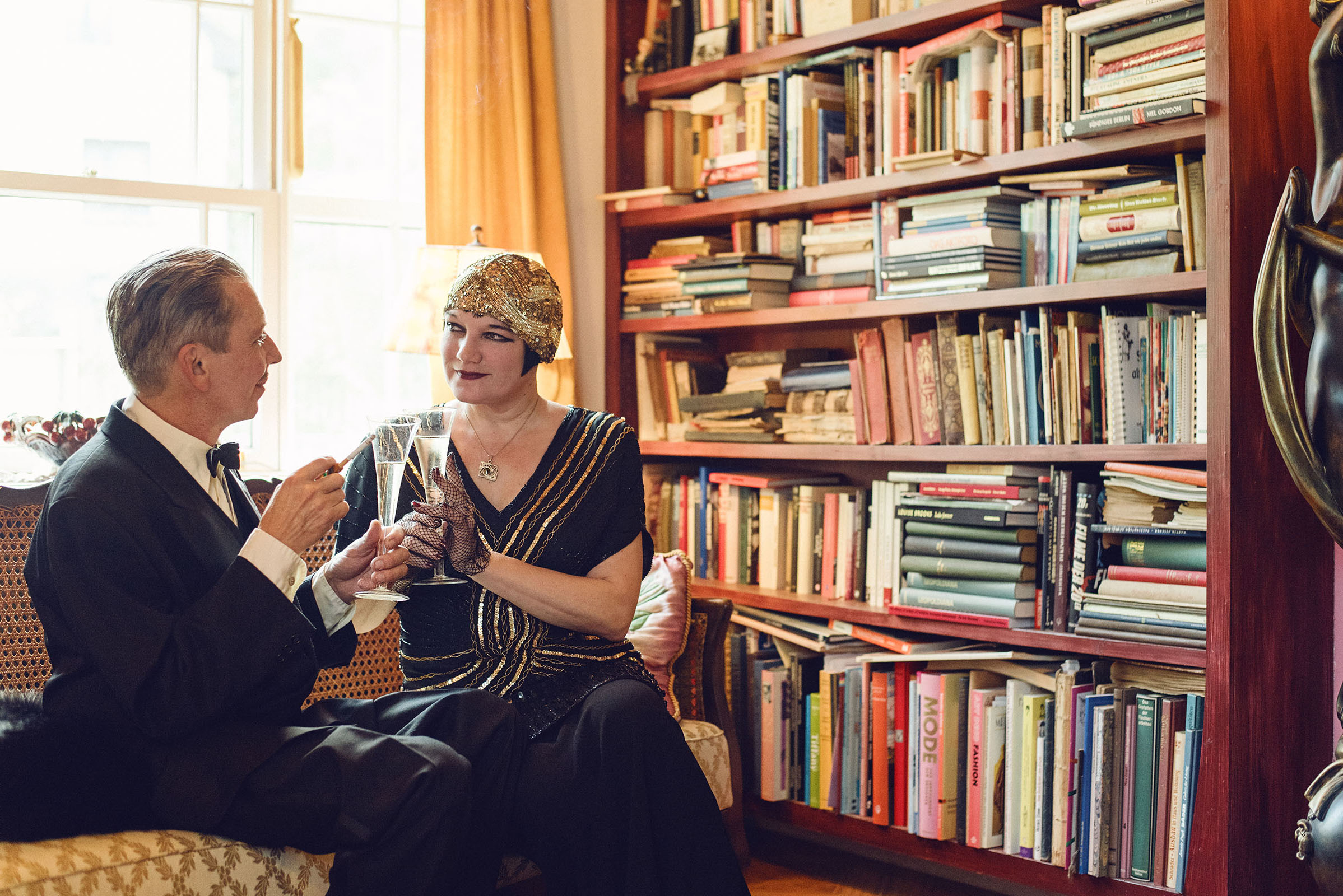 1920s inspired wedding photo shoot by fashion historian and vintage stylist katalina sasdy. a lady and a gentleman are sitting on a yellow sofa in 1920s evening wear and toasting with champagne. the lady is wearing a black silk dress with gold embroidery and a gold headgear. she has a black bob hairstyle and is heavily made up with a cigarette in her hand. the gentleman is wearing a black suit with a white shirt and tie.