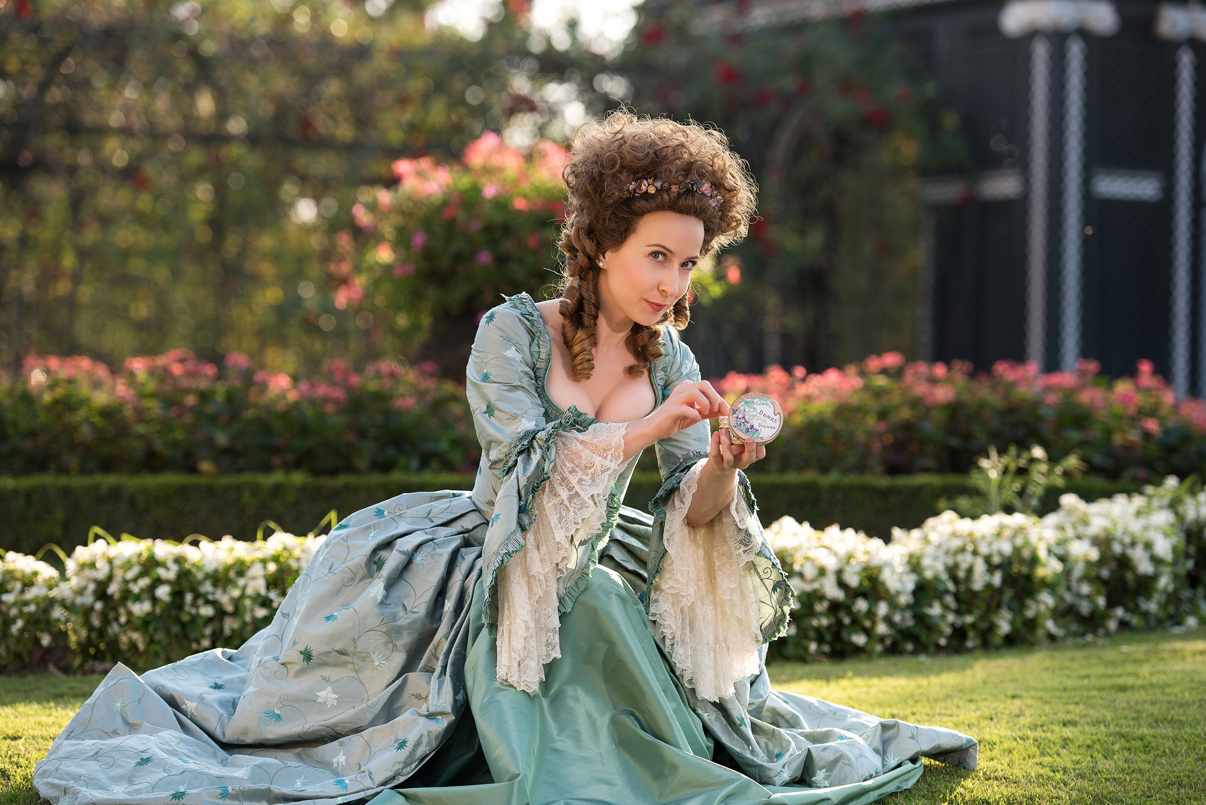 18th century inspired demel advertising photo shoot by fashion stylist katalina sasdy at schoenbrunn palace in vienna. the lady is sitting on the green lawn in the palace garden with a demel candy box in her hands. she is wearing a delicate light turquoise rococo gown with white lace on the sleeves produced by lambert hofer. a we are flowergirls flower ribbon is woven into her brown, curly hedgehog wig.