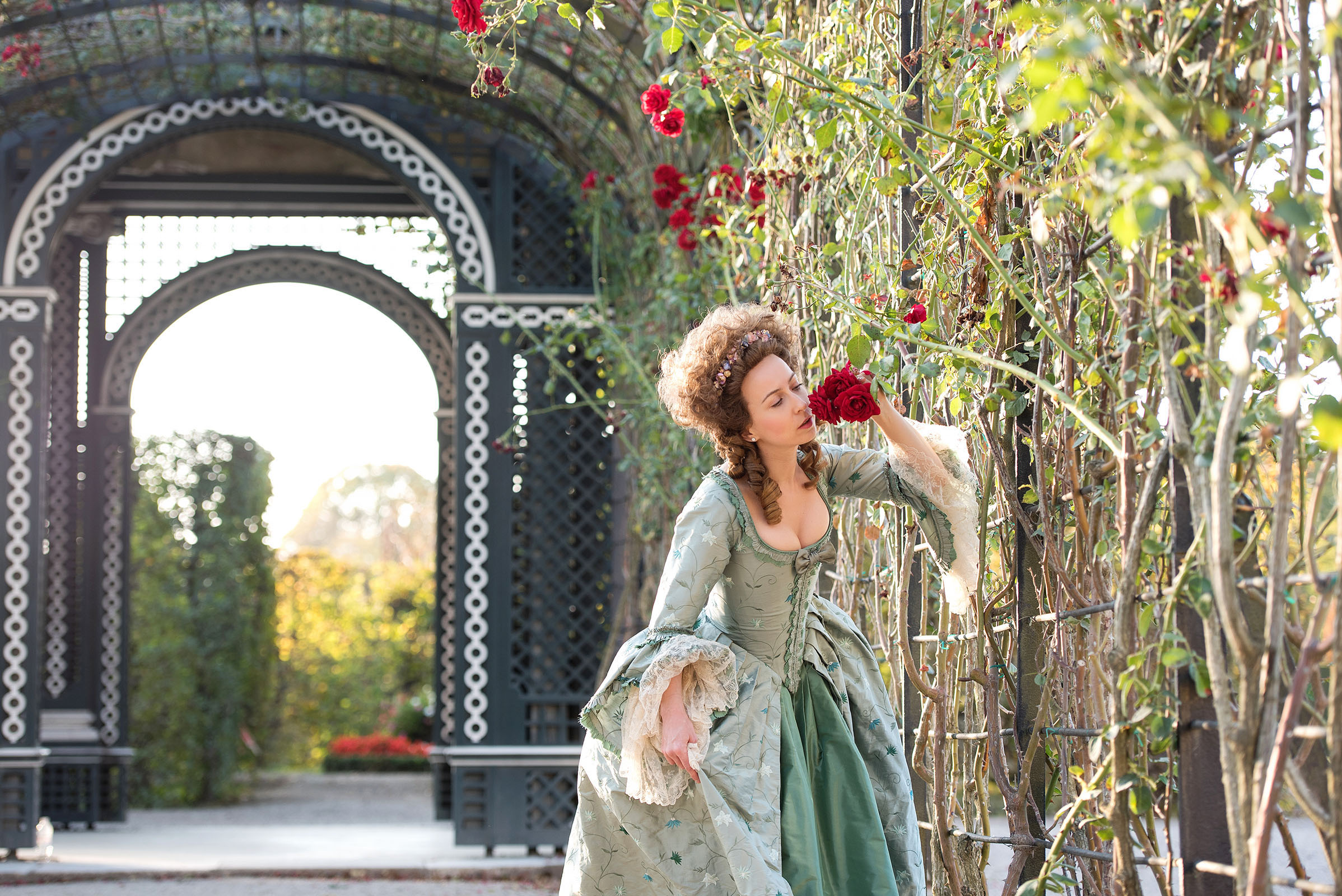 marie antoinette inspired wedding photo shoot by fashion historian and stylist katalina sasdy at schoenbrunn palace in vienna. the lady is standing in the castle garden and smells a red rose. she is wearing a delicate light turquoise rococo gown with white lace on the sleeves produced by lambert hofer. a we are flowergirls flower ribbon is woven into her brown, curly hedgehog wig.