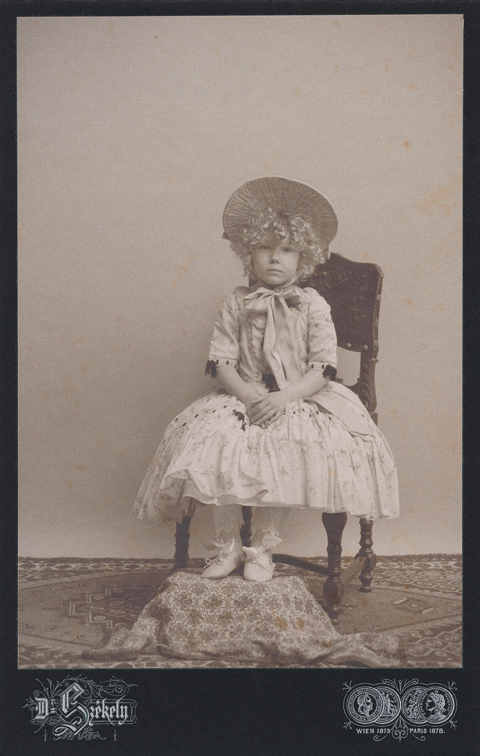 19th century inspired children advertising photo shoot by fashion historian and stylist katalina sasdy. a little girl with blonde curls is sitting on an elaborately decorated armchair in an 1850s dress. her hands are folded together in her lap. she is wearing an elegant hat that is tied under the chin with a bow. her feet, on which she is wearing fine ballerina shoes, are placed on a stool.