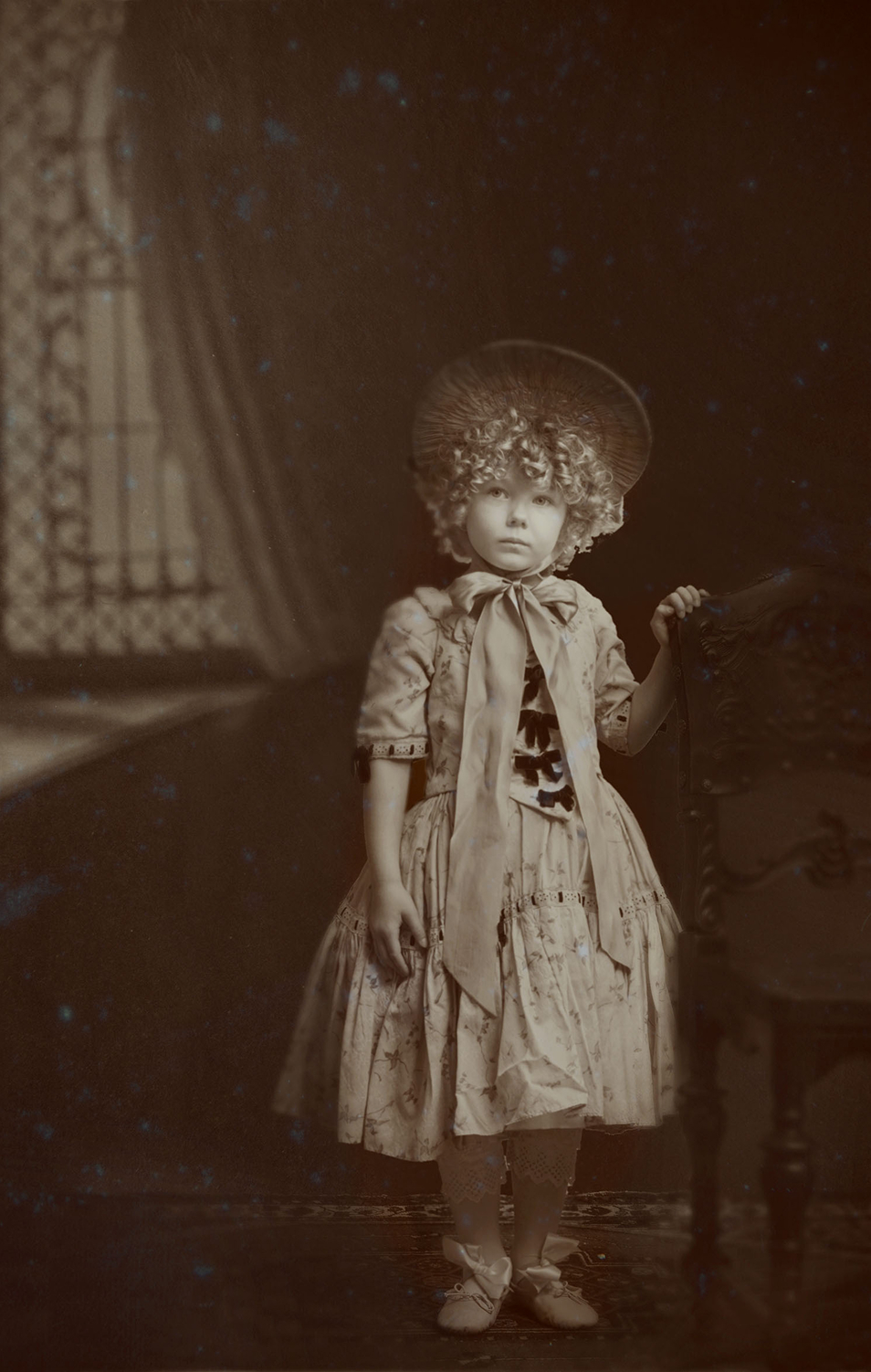 19th century inspired children advertising photo shoot by fashion stylist katalina sasdy. a little girl with blonde curls, wearing a 1850s dress, with an elegant hat that is tied under the chin with a bow, is standing next to an elaborately decorated armchair. her hands are leaning on the back of the chair. on her feets are fine ballerina shoes. in the background there is a half-open curtain.