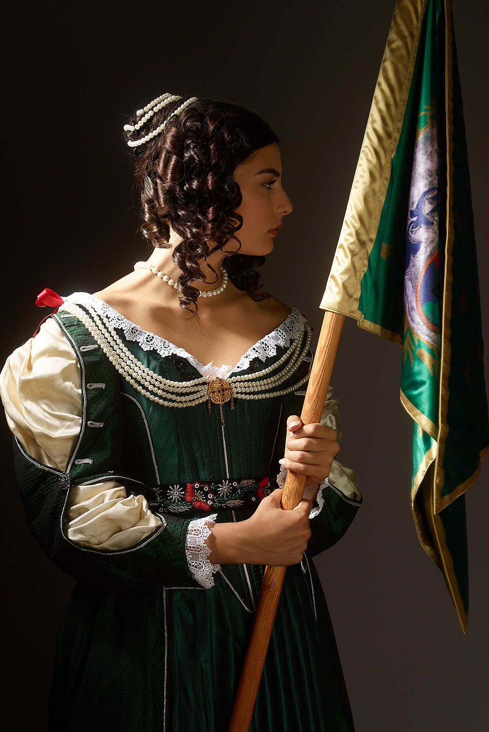 helena zrinski inspired book cover photoshoot by fashion stylist katalina sasdy. helena zrinski is wearing a green silk dress in the style of the 17th century in hungary. the dress is decorated with beads, red ribbons and lace. the parts of the silk blouse are open on her sleeves. she is photographed in profile, so the typical features oft he 17th century hairstyle are visible, a high knot, decorated with pearls and side curls. she carries the flag of her son, the hungarian freedom fighter, prince francis rakoczi II.