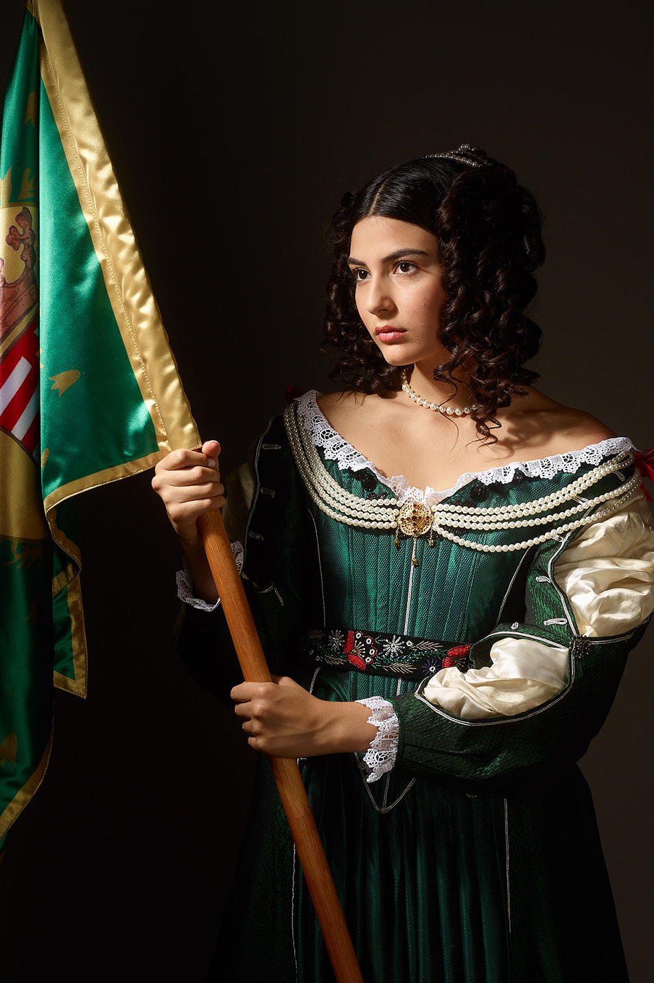 1680s womens fashion inspired advertising photo shoot by art director and fashion stylist katalina sasdy. helena zrinski is wearing a green silk dress in the style of the 17th century in hungary. the dress is decorated with beads, red ribbons and lace. the parts of the silk blouse are open on her sleeves. she carries the flag of her son, the hungarian freedom fighter, prince francis rakoczi II. she is looking dreamily into the distance.