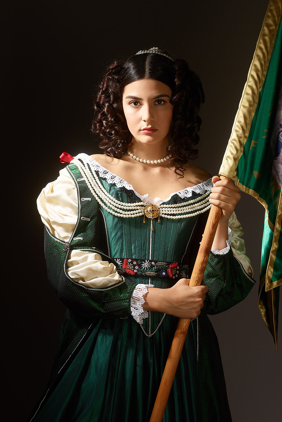 17th century womens fashion inspired advertising photo shoot by fashion historian and stylist katalina sasdy. helena zrinski is wearing a green silk dress in the style of the 17th century in hungary. the dress is decorated with beads, red ribbons and lace. the parts of the silk blouse are open on her sleeves. she carries the flag of her son, the hungarian freedom fighter, prince francis rakoczi II. she is looking resolutely into the camera.