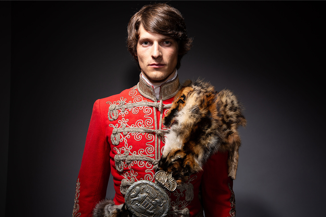 1848 hungarian revolution inspired advertising photo shoot by fashion historian and stylist katalina sasdy. a genteman is wearing a red hussar uniform with a panther skin that is held together with a silver buckle. the uniform includes a belt, braided from silver thread, silver embroidery and uniform decorations. it is a half body picture and the hussar is looking straight into the camera.