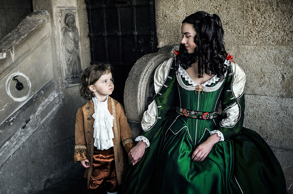 zrinyi ilona and rakoczi ferenc themed advertising photo shoot at the castle of liechtenstein by art director and fashion stylist katalina sasdy. zrinyi ilona is sitting on a stone bench on the terrace of the castle. her little son is standing beside her. they are looking at eachother lovingly. both of them are wearing costumes in stlye of the 17th century. on ilonas green silk dress are lace decorations with red bows on the sleeves and neckline. she is wearing a golden brooch with long pearl necklaces as jewellery. the young prince is wearing a light brown colored silk jacket with cognac colored knee pants and white lace shirt.