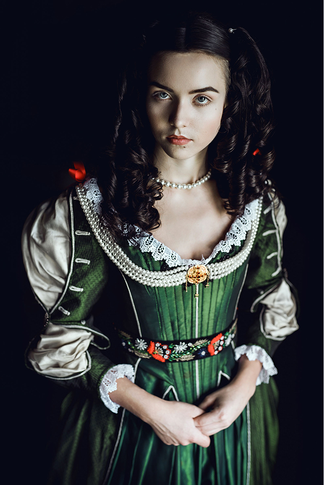 17th century inspired hungarian fashion photoshoot about zrinyi ilona at the castle of liechtenstein by fashion stylist katalina sasdy. zrinyi ilona is looking directly in the camera. she is wearing a costume in the stlye of the 17th century. on her green silk dress are lace decorations with red bows on the sleeves and neckline. she is using a golden brooch with long pearl necklaces as jewellery. her hair is classically styled in the fashion of the 17th century, middle parting with curls on both sides and a bun at the back.