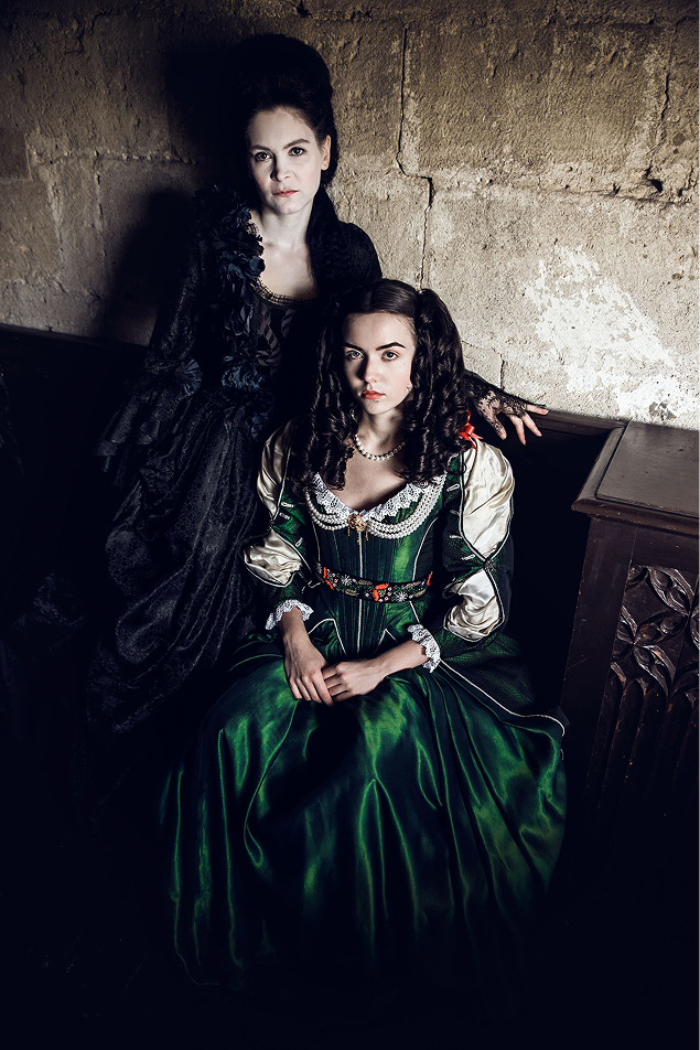 17th - 18th century inspired fashion photo shoot about zrínyi ilona at the castle of liechtenstein by art director and fashion stylist katalina sasdy. zrinyi ilona is sitting on a carved wooden bench with a lady who is dressed entirely in black in the style of the 18th century. zrinyi ilona is wearing a costume in the stlye of the 17th century. on her green silk dress are lace decorations with red bows on the sleeves and neckline. she is using a golden brooch with long pearl necklaces as jewellery. both ladies looking directly into the camera.