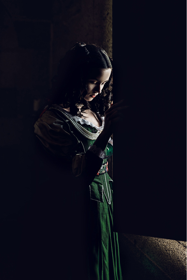 17th century inspired hungarian fashion photo shoot about helena zrinski at the castle of liechtenstein by fashion historian and stylist katalina sasdy. the countess is standing in the dark room and opens a window, which illuminates her face. she is wearing a costume in the stlye of the 17th century. on her green silk dress are lace decorations with red bows on the sleeves and neckline. she is using a golden brooch with long pearl necklaces as jewellery.