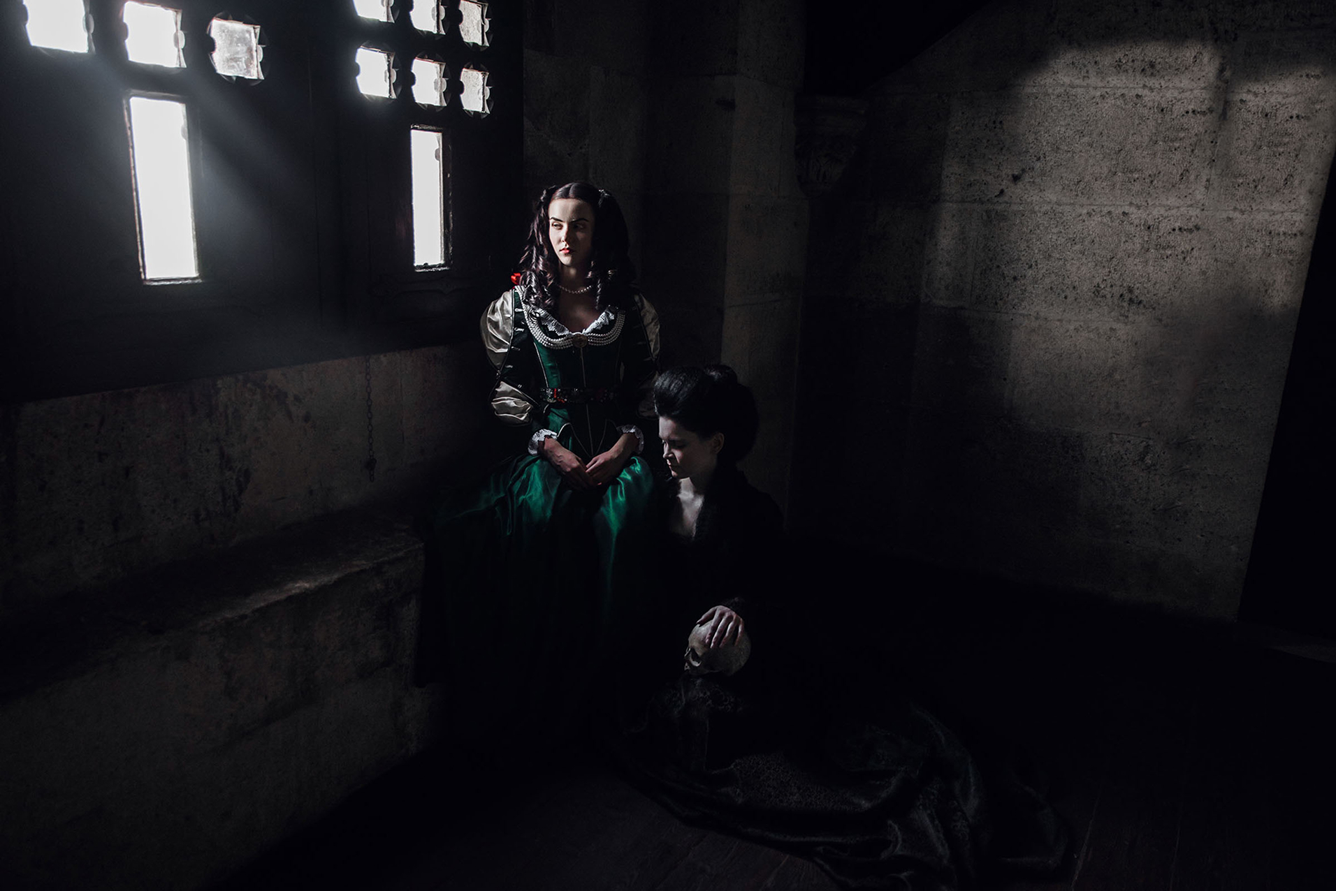 17th - 18th century inspired fashion photo shoot about helena zrinski at the castle of liechtenstein by fashion stylist katalina sasdy. helena zrinski is sitting in a dark prison with a barred window. with her is the lady in the 18th century black dress, who is holding a skull in her hand. the room is completely dark, only the grilled windows let in the light.