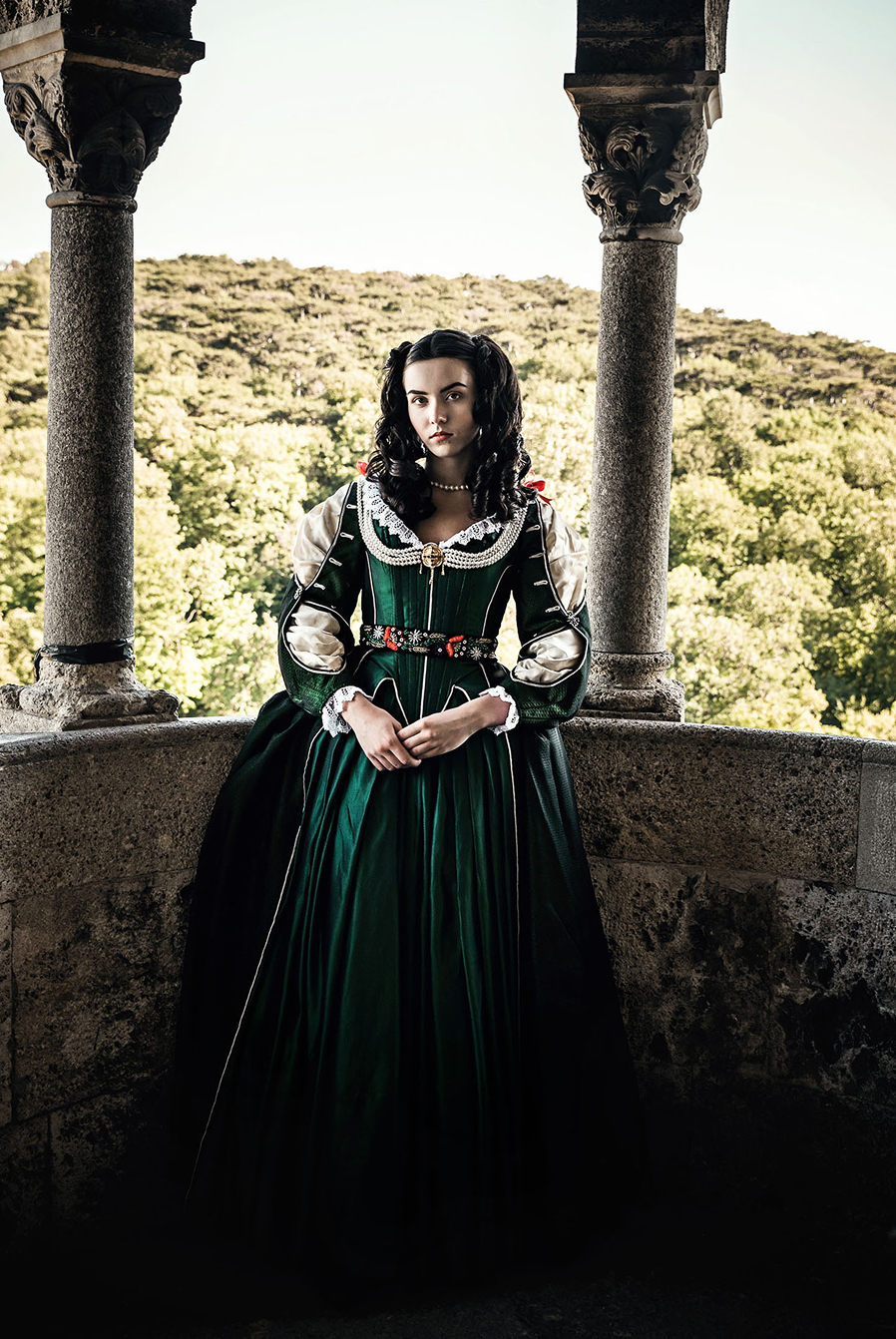 helena zrinski inspired advertising photo shoot at the castle of liechtenstein by fashion stylist katalina sasdy. helena zrinski is standing on the pillared balcony of the castle in a green silk dress in the fashion of the 17th century. on the dress are lace decorations with red bows on the sleeves and neckline. she is wearing a golden brooch with long pearl necklaces as jewellery.
