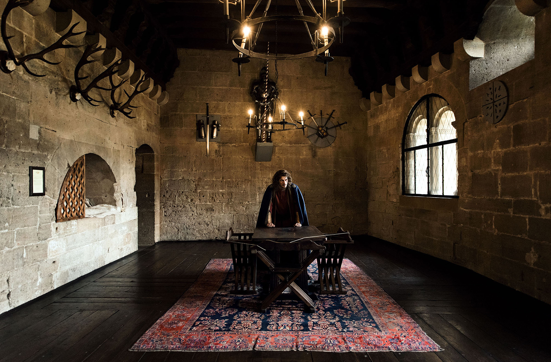advertising photoshoot of the royals of liechtenstein by fashion historian and stylist katalina sasdy. the prince of liechtenstein is standing on a colorful carpet in the middle of the large banquet hall with his fists leaning on a table. there is a lampstand above him. he is wearing the aristocratic fashion of the 12th century, a red linen tunic, which sleeves are richly embroidered with gold thread and a dark blue cape, fastened at the shoulder with gold rivets. the princes light brown hair is shoulder-length and wavy.