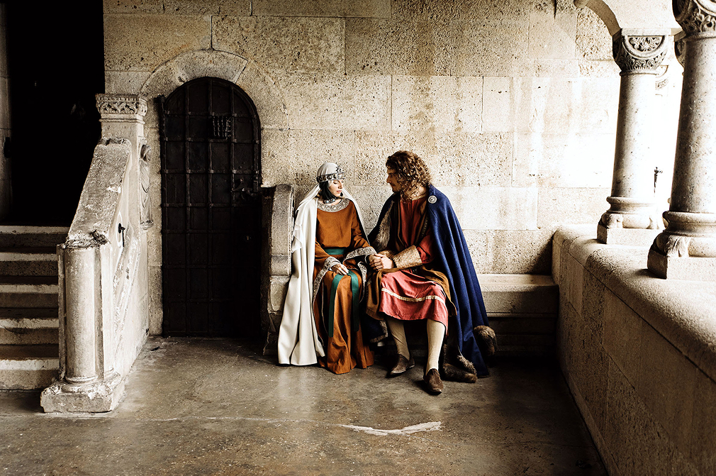 middle age inspired wedding photo shoot in the castle of liechtenstein by art director and fashion stylist katalina sasdy. the prince of liechtenstein is sitting hand in hand on the castle terrace with his wife. both are dressing in the aristocratic fashion of the 12th century. the prince is wearing a red linen tunic, which sleeves are richly embroidered with gold thread, light green stockings and a dark blue cape, fastened at the shoulder with gold rivets. the shoes are made of brown leather. his light brown hair is shoulder-length and wavy. the princess of liechtenstein is dressing in a long brown gown with bronze embroidery and a green waist belt. as headwear, she is wearing a silver tiara tied under her chin with dark green ribbons and a long white veil. her hair is braided into two long braids. she gazing shyly at the floor while her husband is looking at her lovingly.