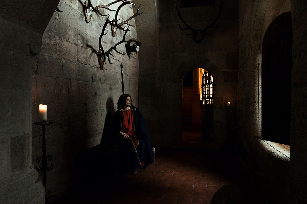 12th-century-inspired-advertising-photo shoot in the castle of liechtenstein by fashion historian and stylist katalina sasdy. the prince of liechtenstein is sitting thoughtfully in a dark room in his castle. only an open window provides light. there are deer antlers on the walls. the prince is wearing the aristocratic fashion of the 12th century, a red linen tunic, which sleeves are richly embroidered with gold thread and a dark blue cape, fastened at the shoulder with gold rivets. the princes light brown hair is shoulder-length and wavy.