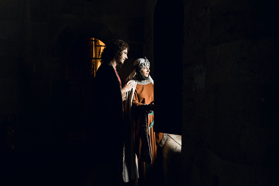 robin hood wedding inspiration photo shoot in the castle of liechtenstein by art director and fashion stylist katalina sasdy. the prince and princess of liechtenstein are standing in a dark room in front of the open window and are looking outside. both are dressing in the aristocratic fashion of the 12th century. the prince is wearing a red linen tunic, which sleeves are richly embroidered with gold thread, light green stockings and a dark blue cape, fastened at the shoulder with gold rivets. his light brown hair is shoulder-length and wavy. the princess of liechtenstein is dressing in a long brown gown with bronze embroidery and a green waist belt. as headwear, she is wearing a silver tiara tied under her chin with dark green ribbons and a long white veil. her hair is braided into two long braids. 