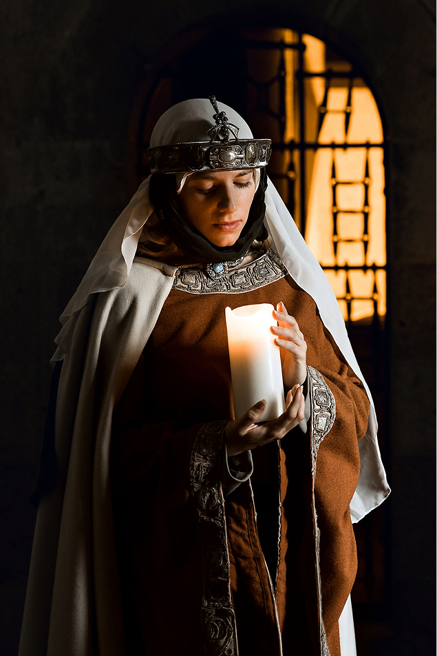 advertising photo shoot of the princess of liechtenstein by fashion historian and stylist katalina sasdy. the princess of liechtenstein is standing in a dark room with a candle in her hand. in the background there is a half open gate that is fully illuminated. the princess of liechtenstein is dressing in a long brown gown with bronze embroidery and a green waist belt. as headwear, she is wearing a silver tiara tied under her chin with dark green ribbons and a long white veil. her hair is braided into two long braids.