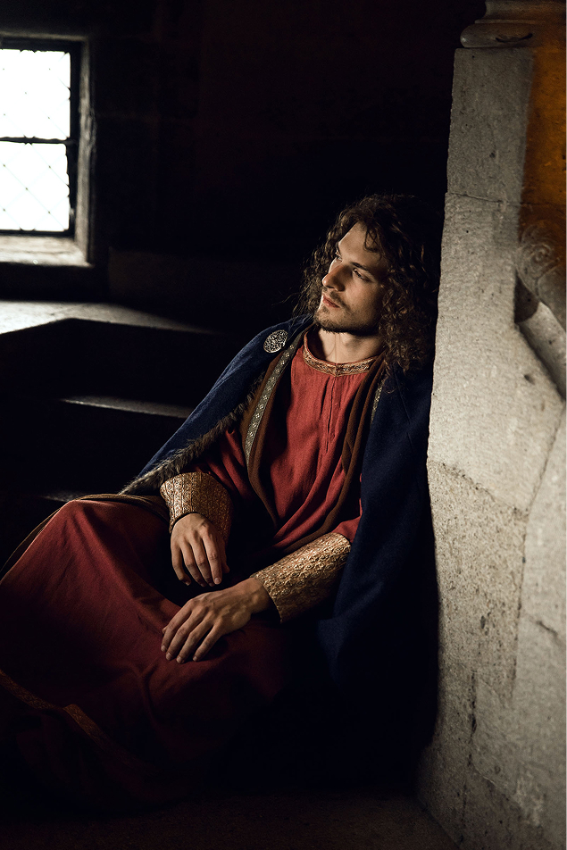 advertising photo shoot of the prince of liechtenstein by fashion stylist katalina sasdy. the prince of liechtenstein is sitting in a dark staircase in the castle and is looking towards the window where the light comes in. the prince is dressing in the aristocratic fashion of the 12th century. he is wearing a red linen tunic, which sleeves are richly embroidered with gold thread, light green stockings and a dark blue cape, fastened at the shoulder with gold rivets. the shoes are made of brown leather. his light brown hair is shoulder-length and wavy.