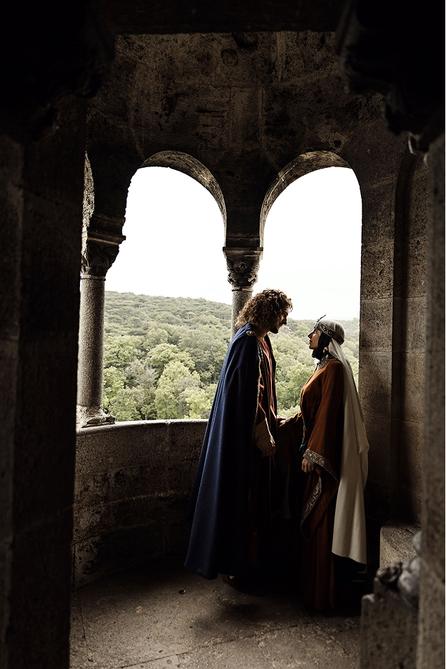 middle age inspired wedding photo shoot in the castle of liechtenstein by art director and fashion stylist katalina sasdy. the prince and princess of liechtenstein are standing on a terrace of the castle surrounded by columns and are looking at each other in love. both are dressing in the aristocratic fashion of the 12th century. the prince is wearing a red linen tunic, which sleeves are richly embroidered with gold thread, light green stockings and a dark blue cape, fastened at the shoulder with gold rivets. the shoes are made of brown leather. his light brown hair is shoulder-length and wavy. the princess of liechtenstein is dressing in a long brown gown with bronze embroidery and a green waist belt. as headwear, she is wearing a silver tiara tied under her chin with dark green ribbons and a long white veil. her hair is braided into two long braids.