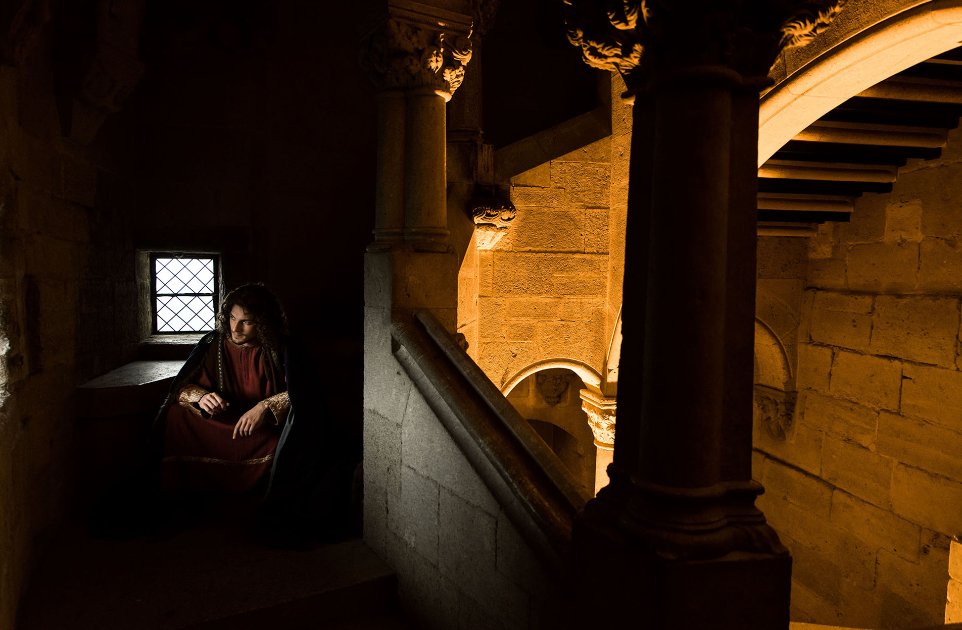 advertising photo shoot of the prince of liechtenstein by fashion historian and stylist katalina sasdy. the prince of liechtenstein is sitting in a dark staircase in the castle and is looking towards the window where the light comes in. the staircase on the right is brightly lit. he is dressing in the aristocratic fashion of the 12th century. he is wearing a red linen tunic, which sleeves are richly embroidered with gold thread, light green stockings and a dark blue cape, fastened at the shoulder with gold rivets. the shoes are made of brown leather. his light brown hair is shoulder-length and wavy.