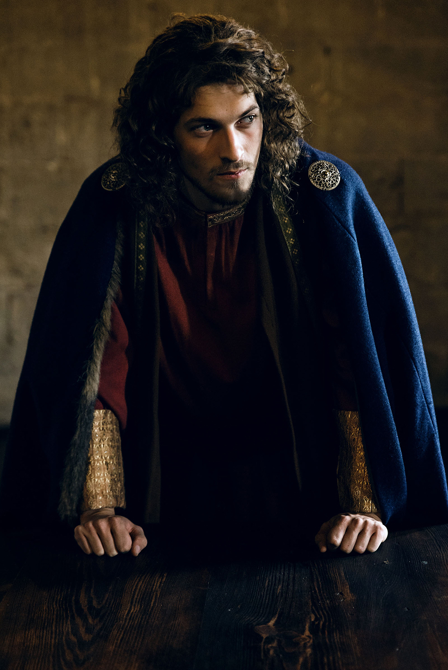 advertising photo shoot of the prince of liechtenstein by fashion stylist katalina sasdy. the prince of liechtenstein is leaning on a table with his fists. he is wearing the aristocratic fashion of the 12th century, a red linen tunic, which sleeves are richly embroidered with gold thread and a dark blue cape, fastened at the shoulder with gold rivets. the princes light brown hair is shoulder-length and wavy.