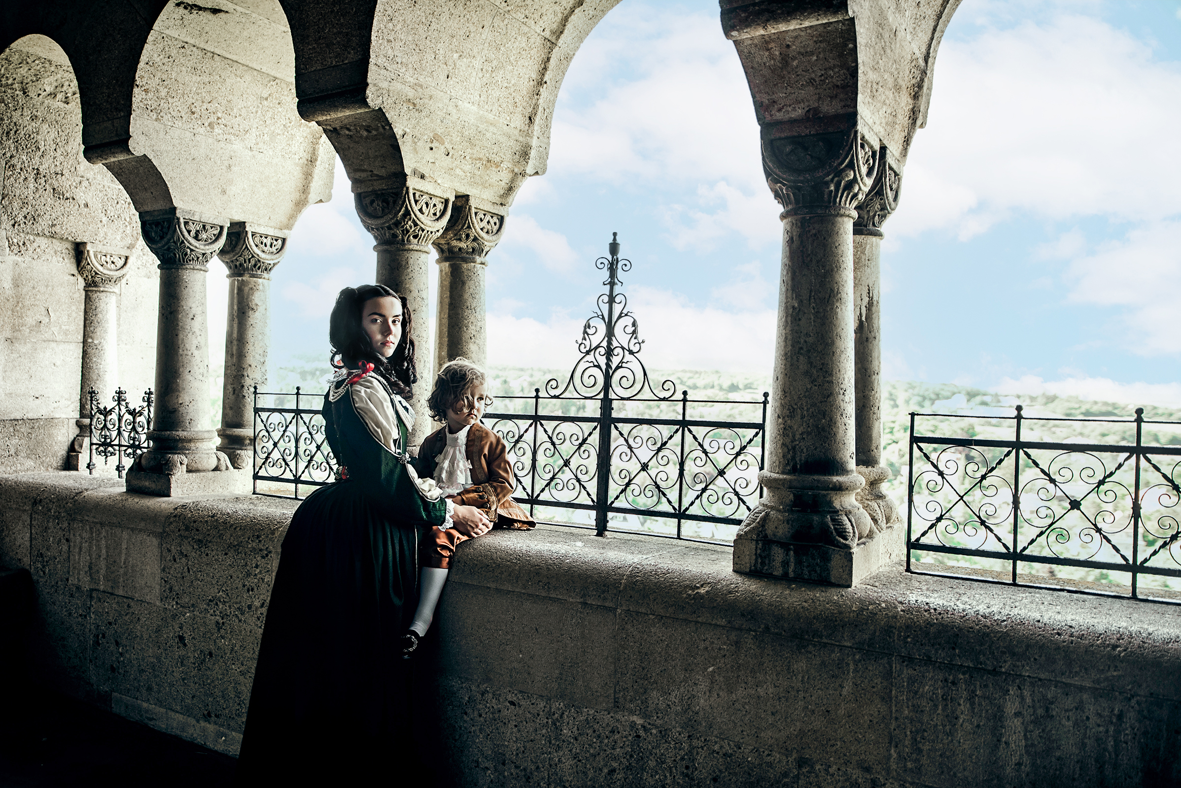 helena zrinski and francis rakoczi inspired advertising photo shoot at the castle of liechtenstein by fashion historian and stylist katalina sasdy. helena zrinski is standing on the balcony of the castle and turning to the camera. her little son, francis rakoczi is sitting on a beam and looking at his mother with interest. both of them wearing costumes in stlye of the 17th century. on helenas green silk dress are lace decorations with red bows on the sleeves and neckline. she is wearing a golden brooch with long pearl necklaces as jewellery. the young prince is wearing a light brown colored silk jacket with cognac colored knee pants and white lace shirt.